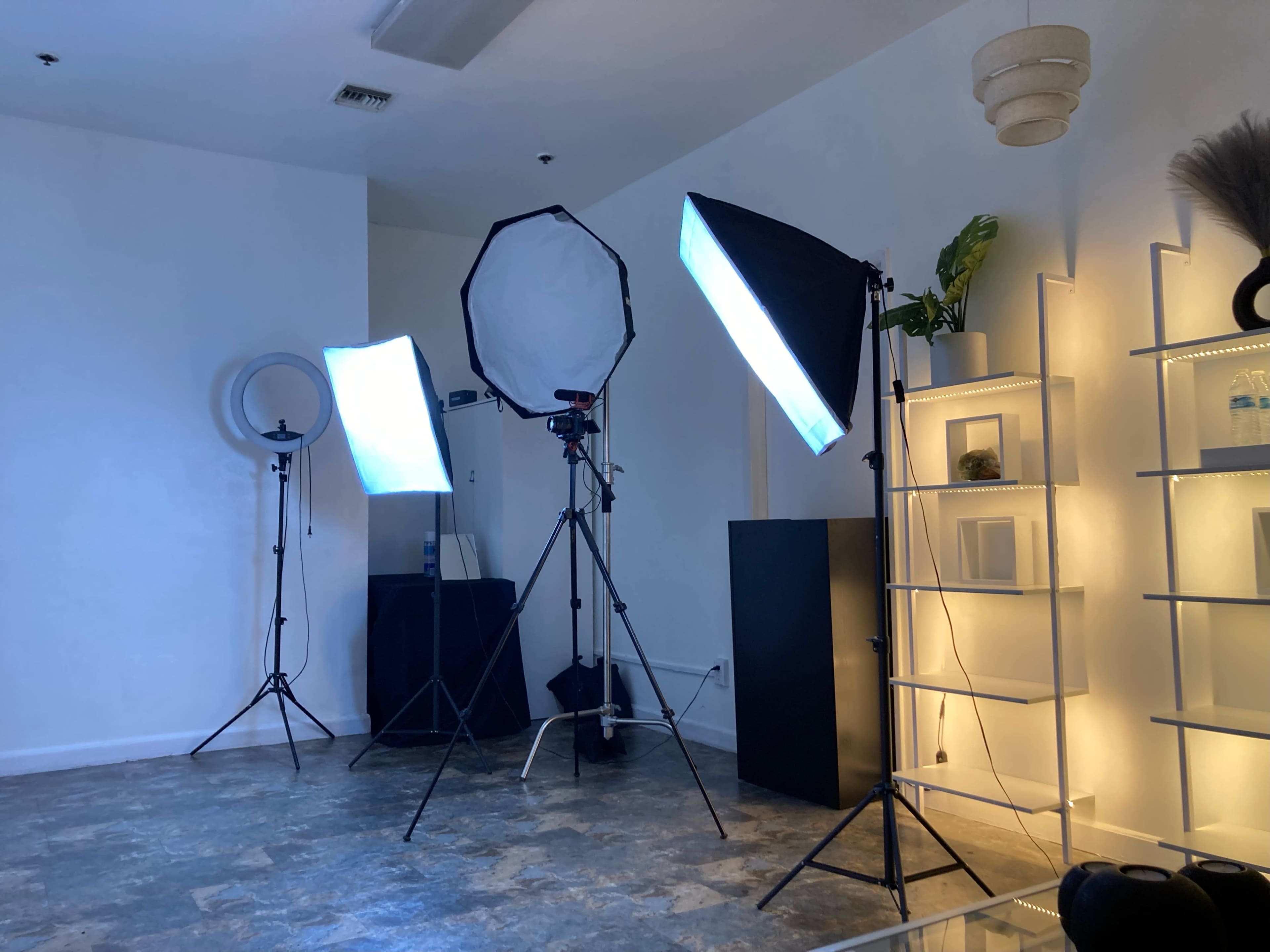 A photography studio setup with three light stands, including a softbox and a ring light, next to a shelving unit and a black cabinet.