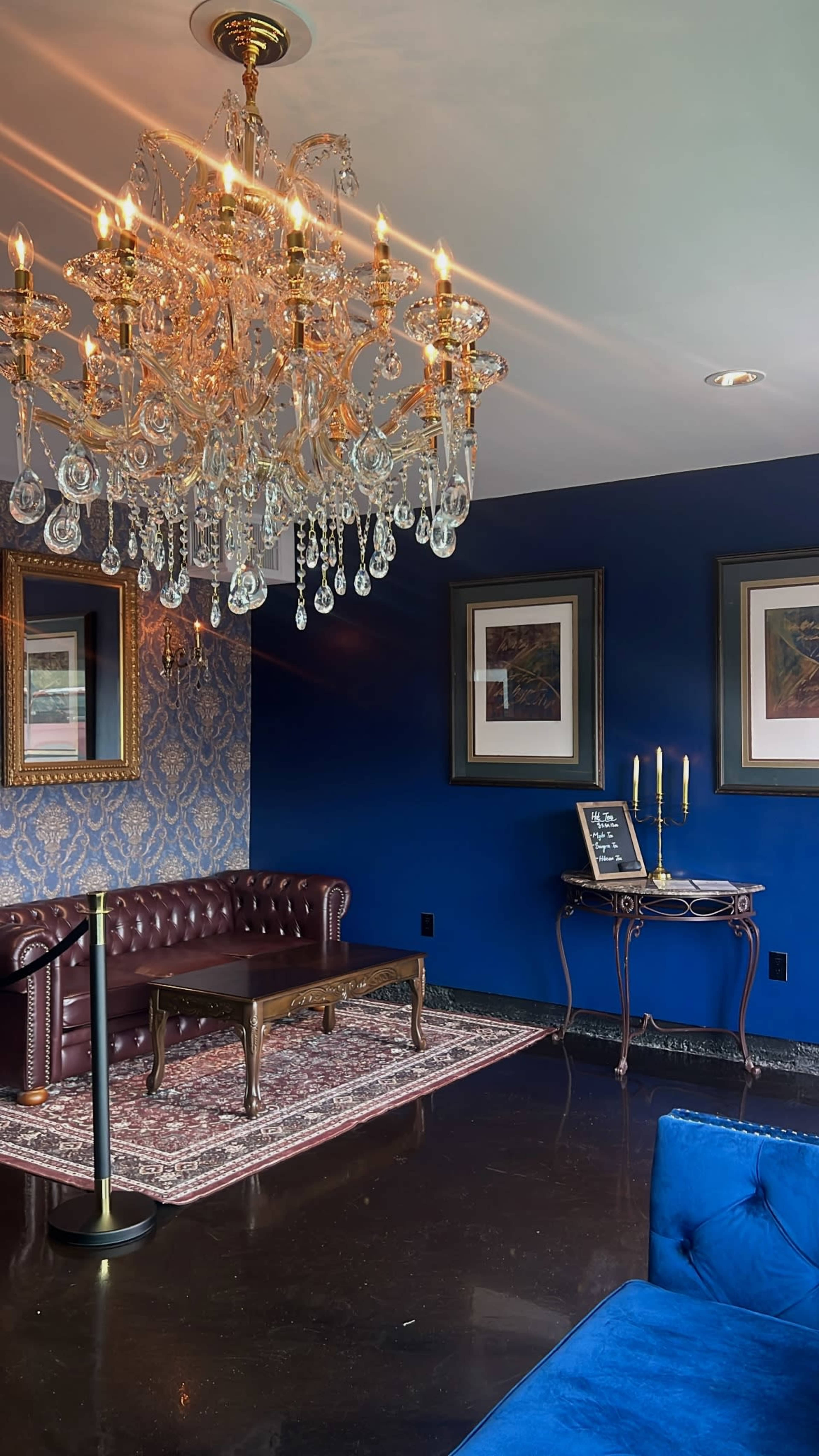 The image shows a luxurious room featuring a chandelier, a burgundy leather couch, a wooden coffee table, and decorative artwork against a dark blue wall.