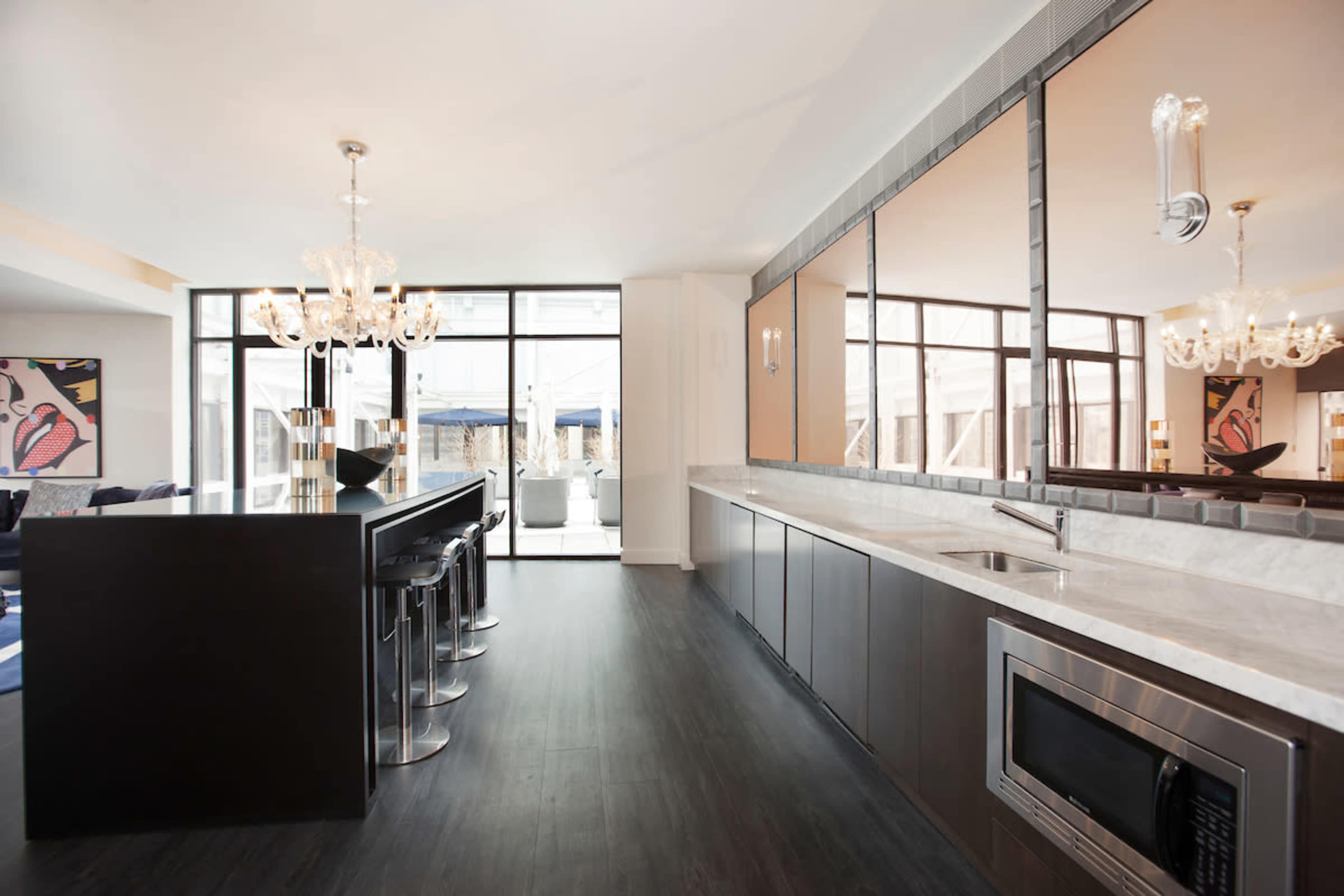 The image shows a modern kitchen with a bar counter, high stools, and large windows allowing natural light to enter the space.