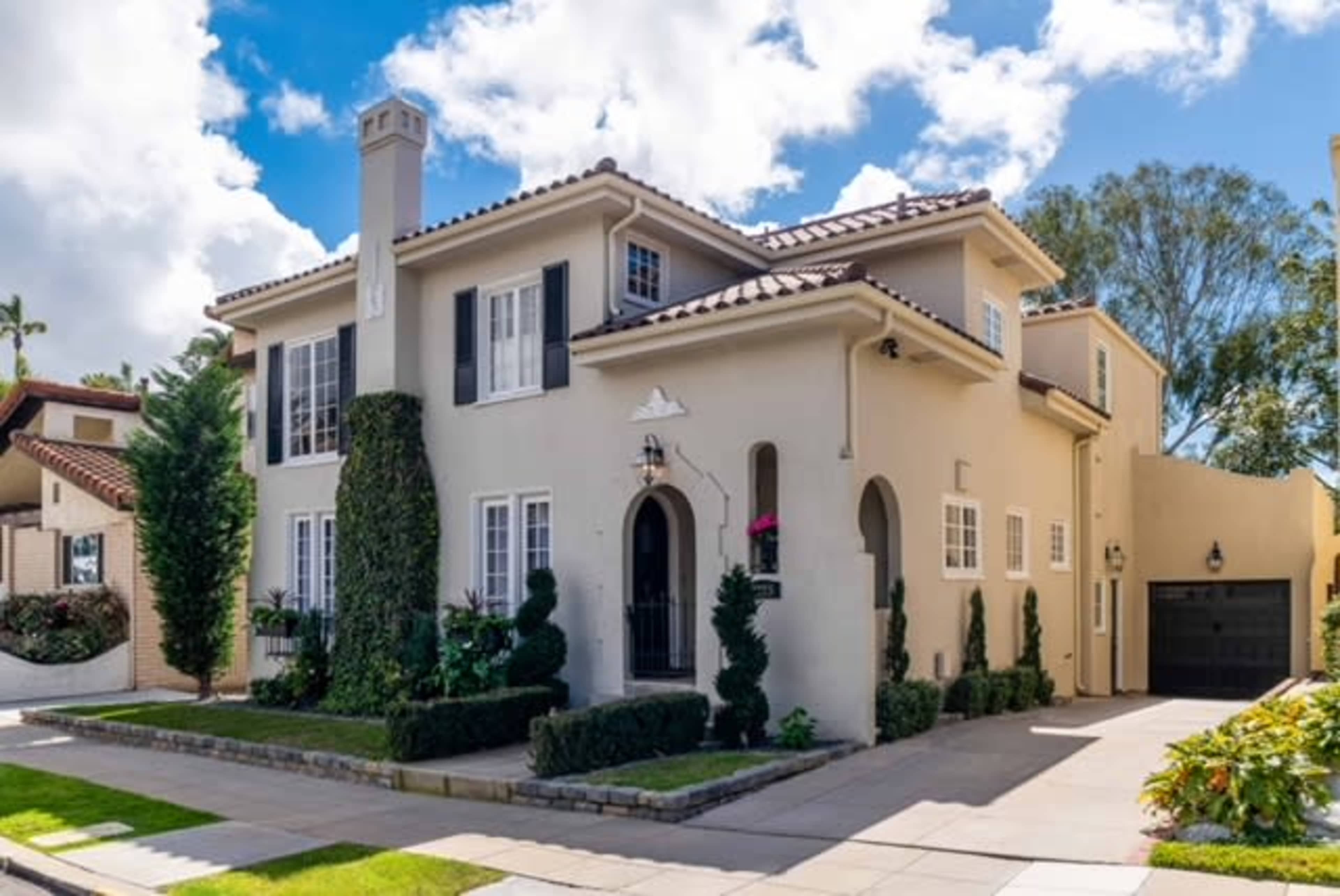 A two-story house with a cream-colored exterior features a tile roof, neatly trimmed hedges, and a garage on the side.