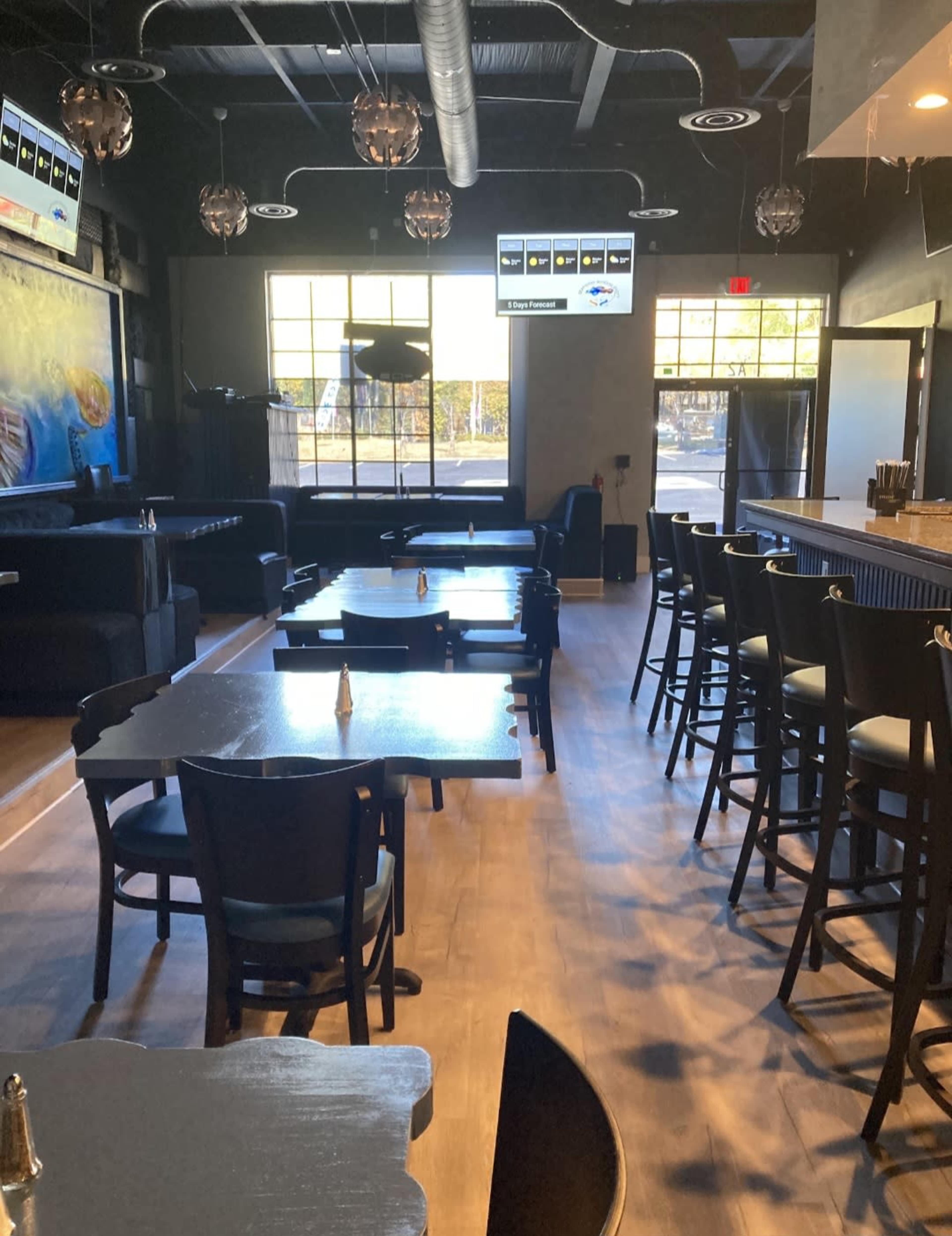 The interior of a restaurant features black tables and chairs arranged in rows, with a bar area and large windows allowing natural light to fill the space.
