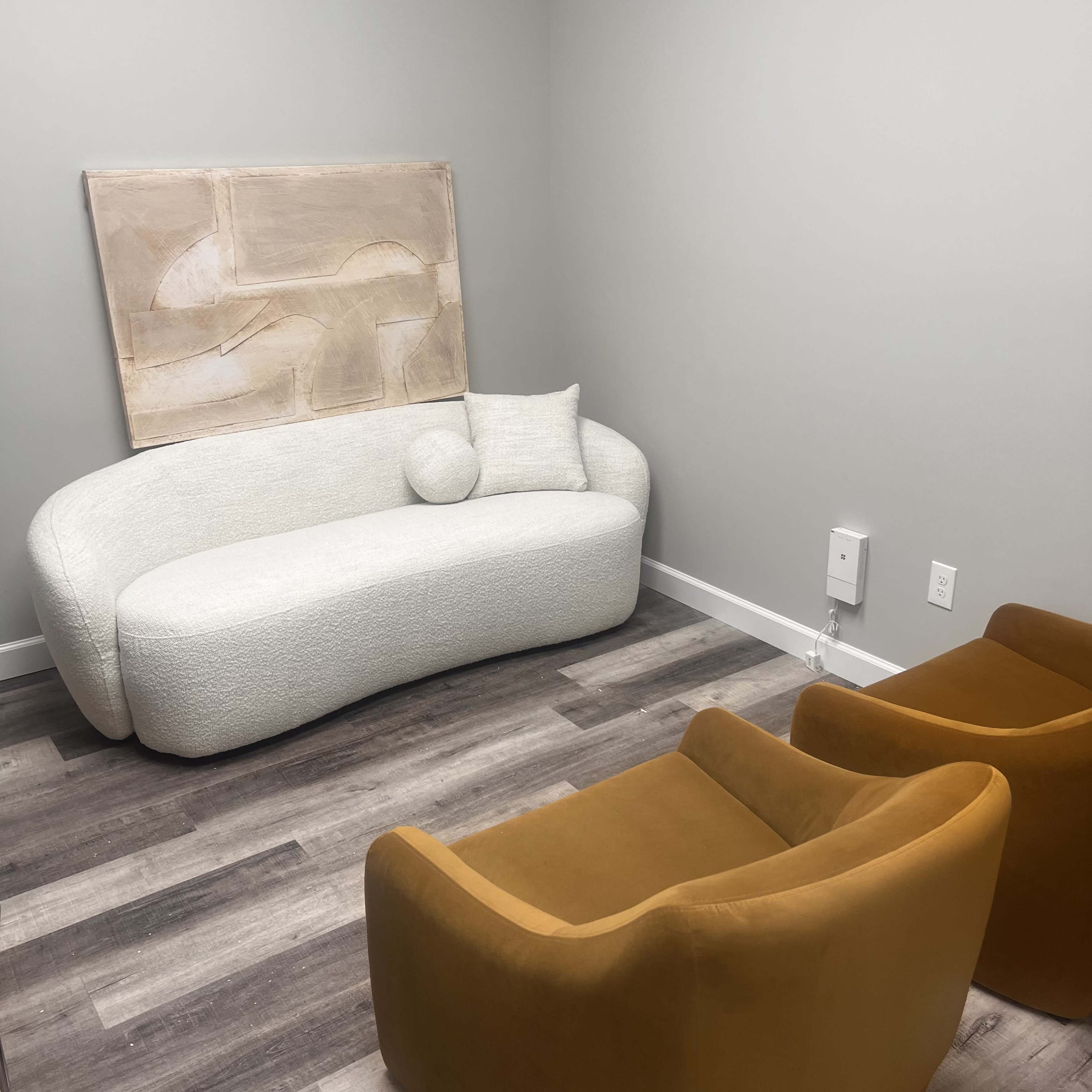 The image shows a small room featuring a white curved couch, a round pillow, two orange armchairs, and an abstract artwork on the wall.