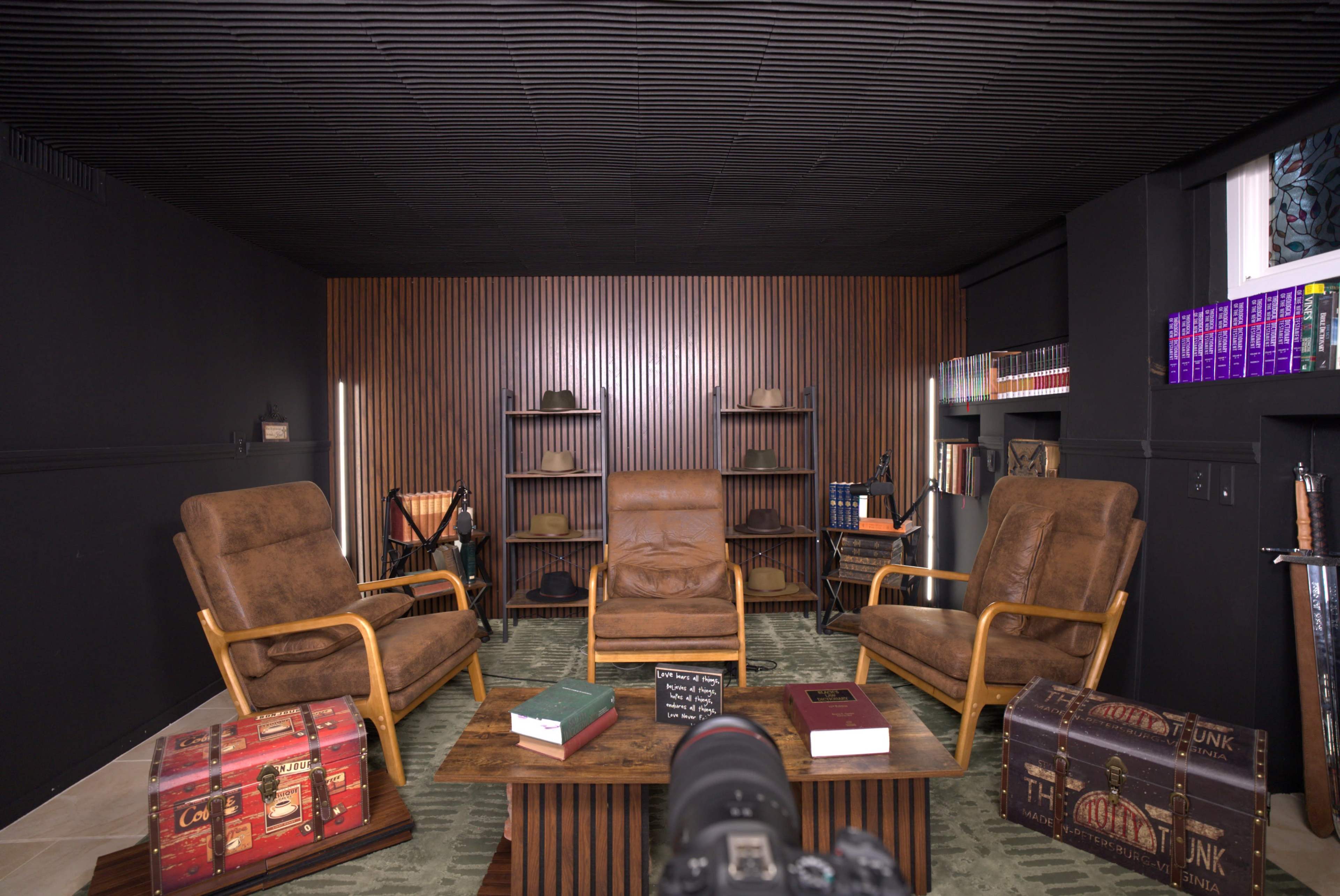 The image depicts a dimly lit room with three brown armchairs surrounding a wooden coffee table, flanked by shelves filled with books and decorative items.