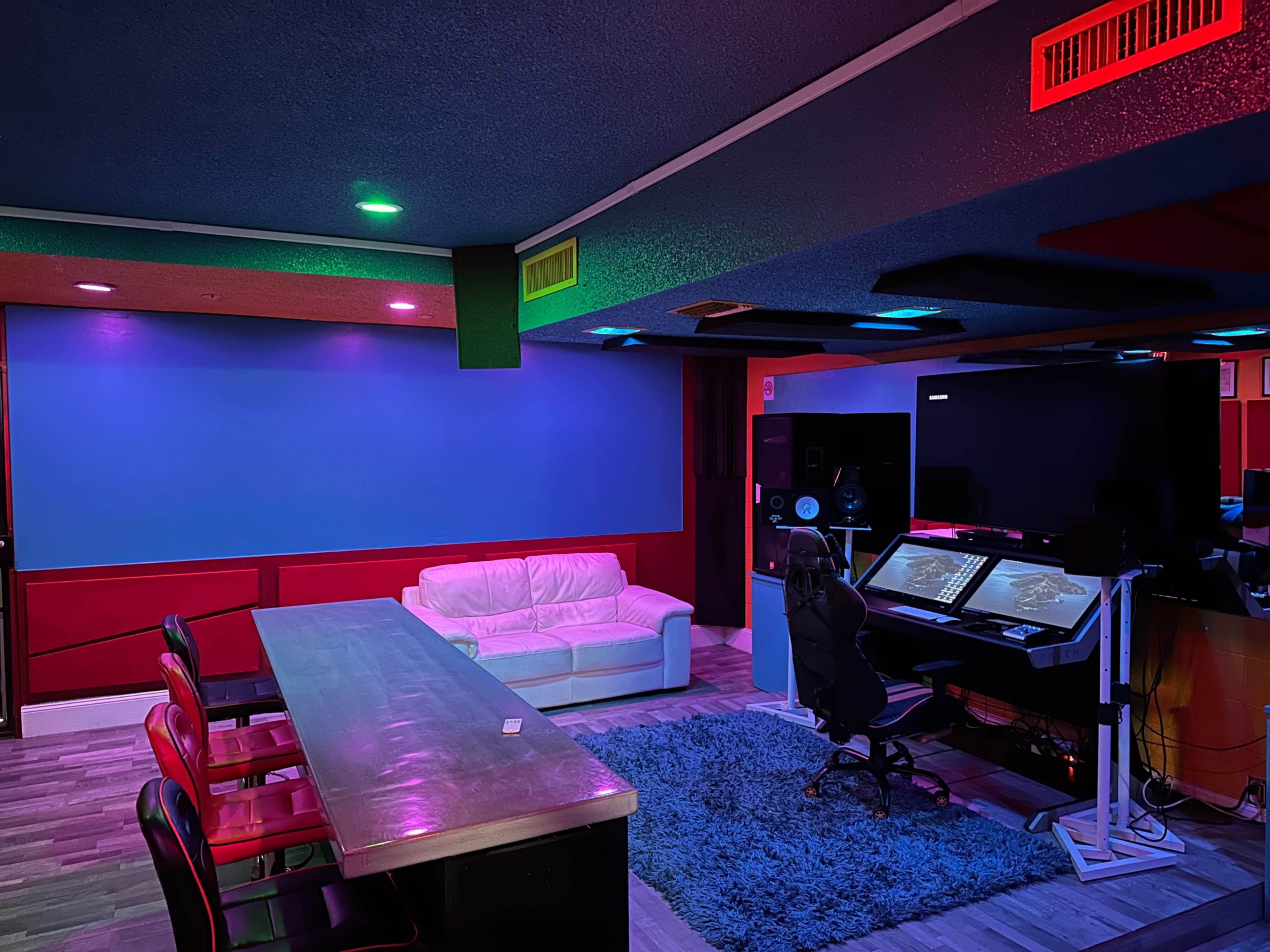 The image shows a modern recording studio or workspace featuring a white couch, a long table with chairs, and a large monitor setup against colorful, illuminated walls.
