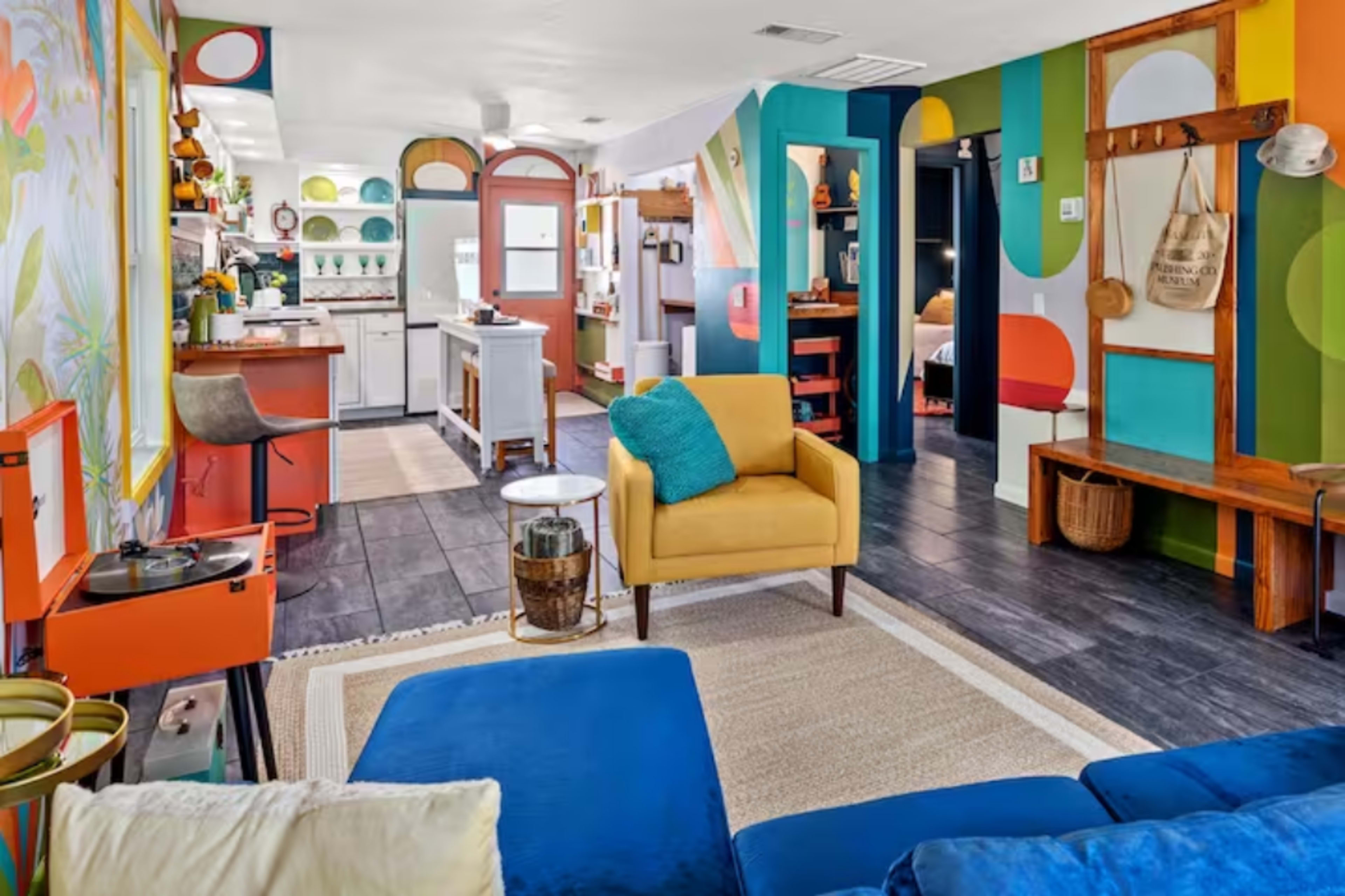 A brightly decorated living space featuring a yellow armchair, a blue sofa, and colorful wall patterns, with a kitchen area visible in the background.