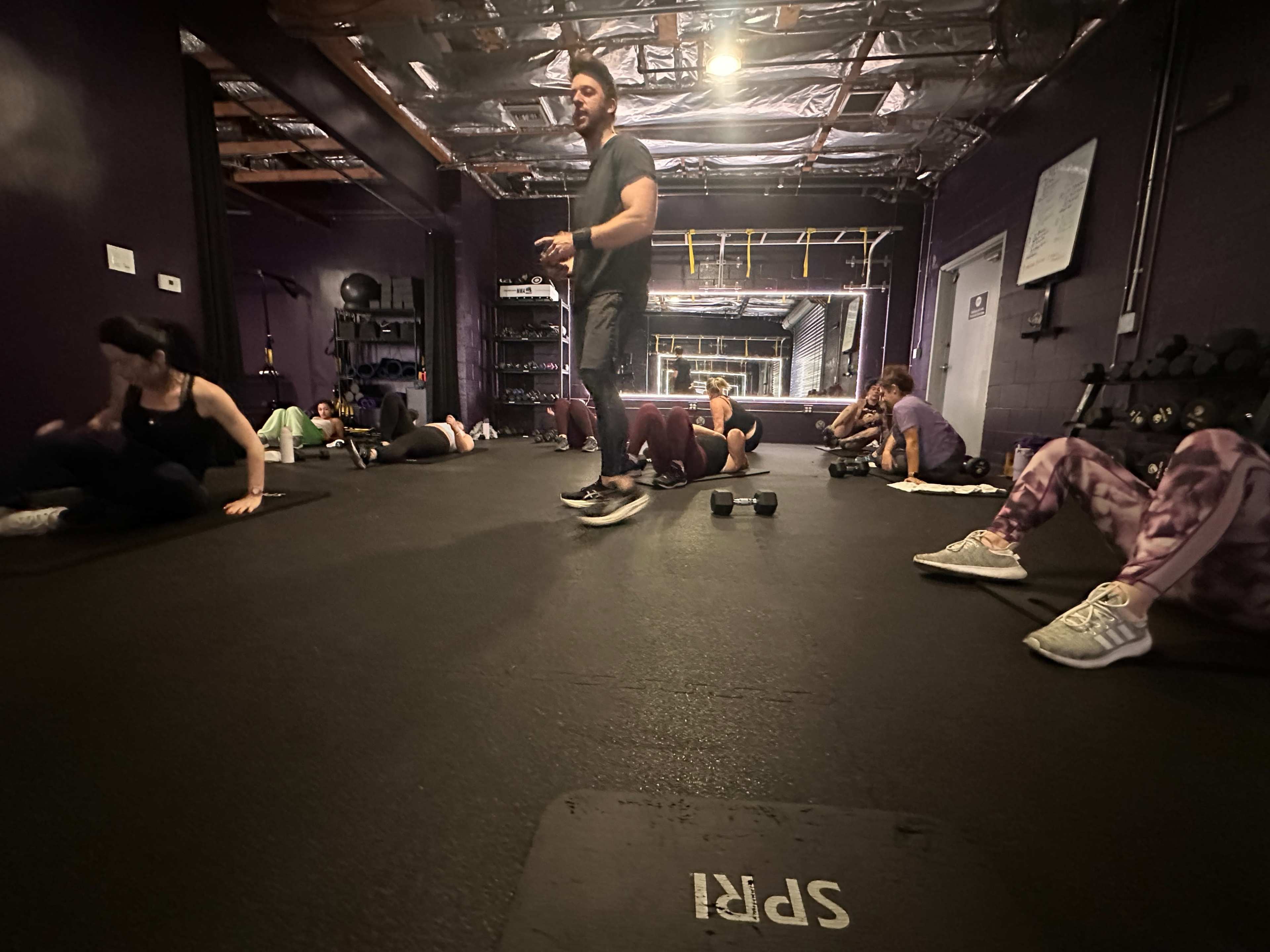 A fitness instructor leads a workout session with participants on the floor, some exercising while others rest in a gym setting.