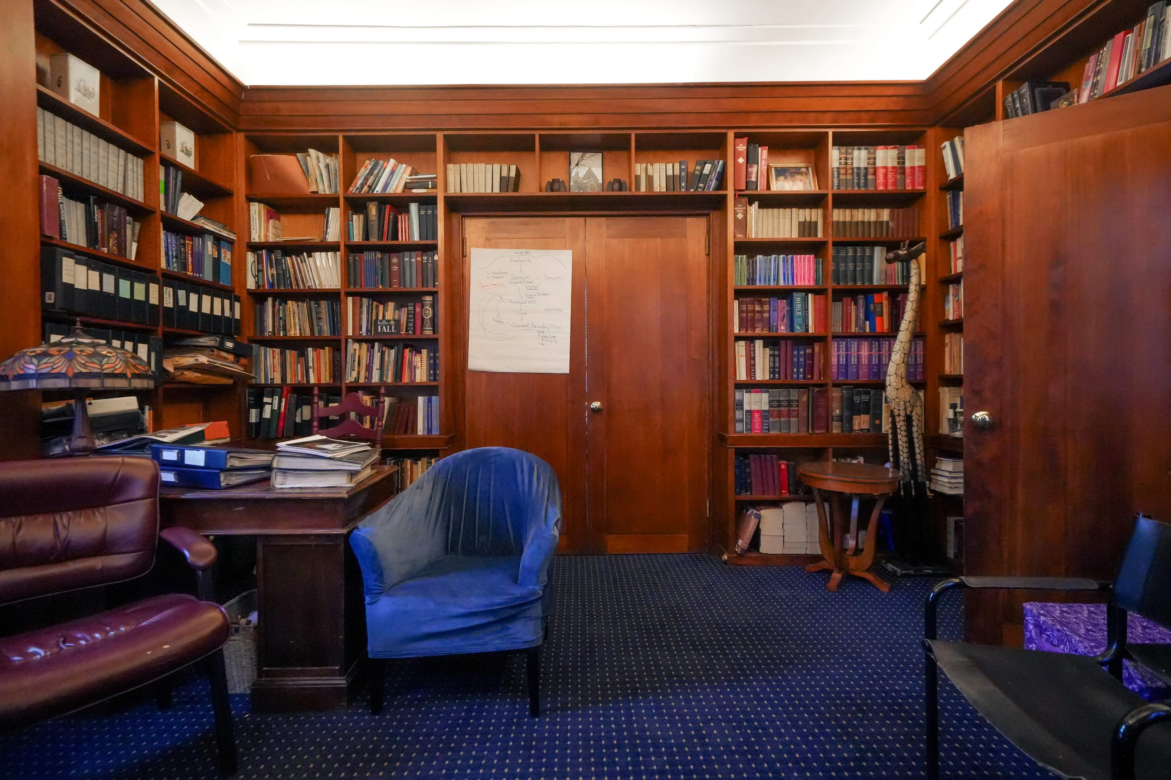 A wooden-paneled library with bookshelves filled with books, a dark blue chair, a desk, and a door leading to another room.