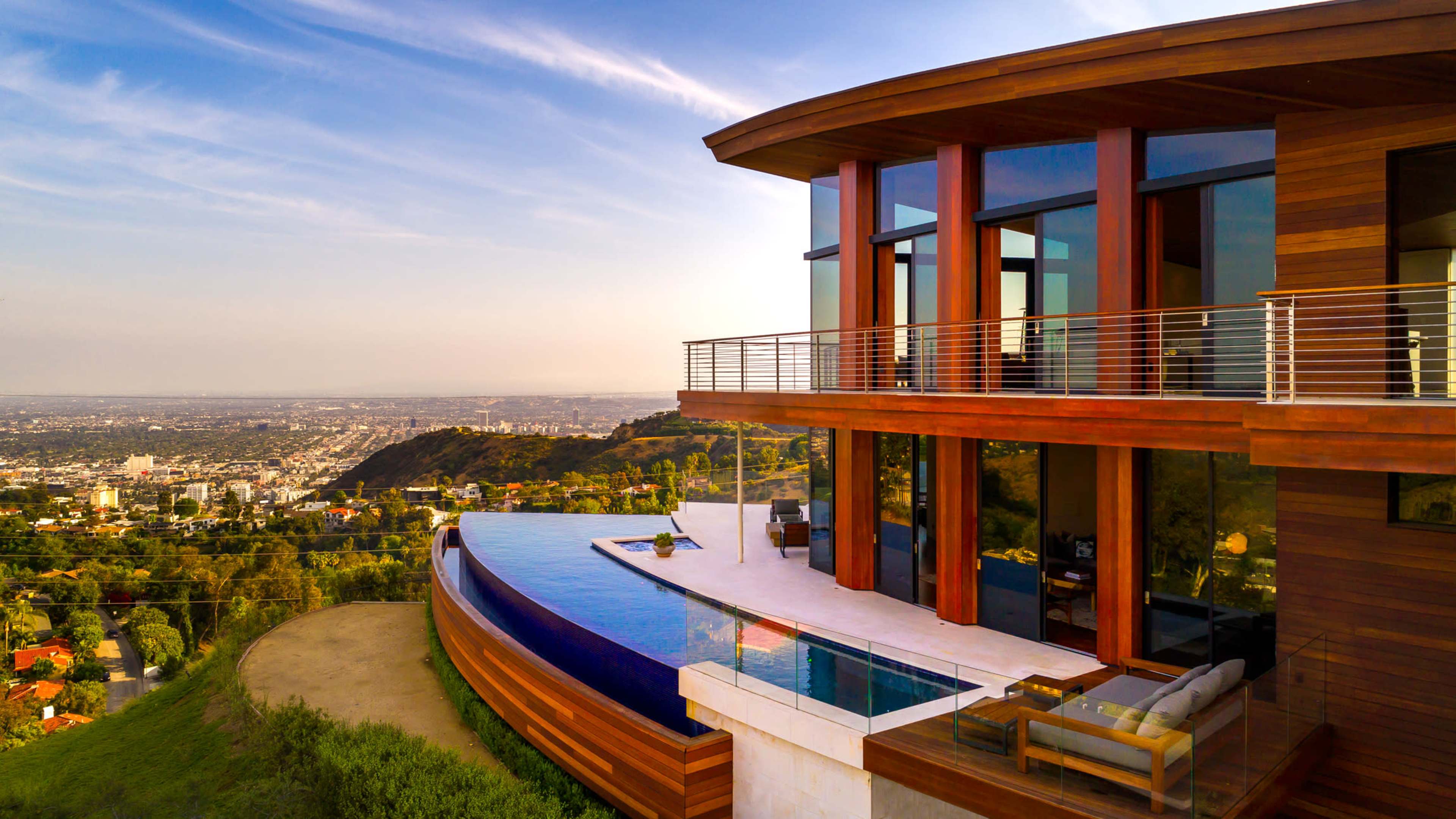 A modern hillside house features large glass windows and an infinity pool that overlooks a city skyline.