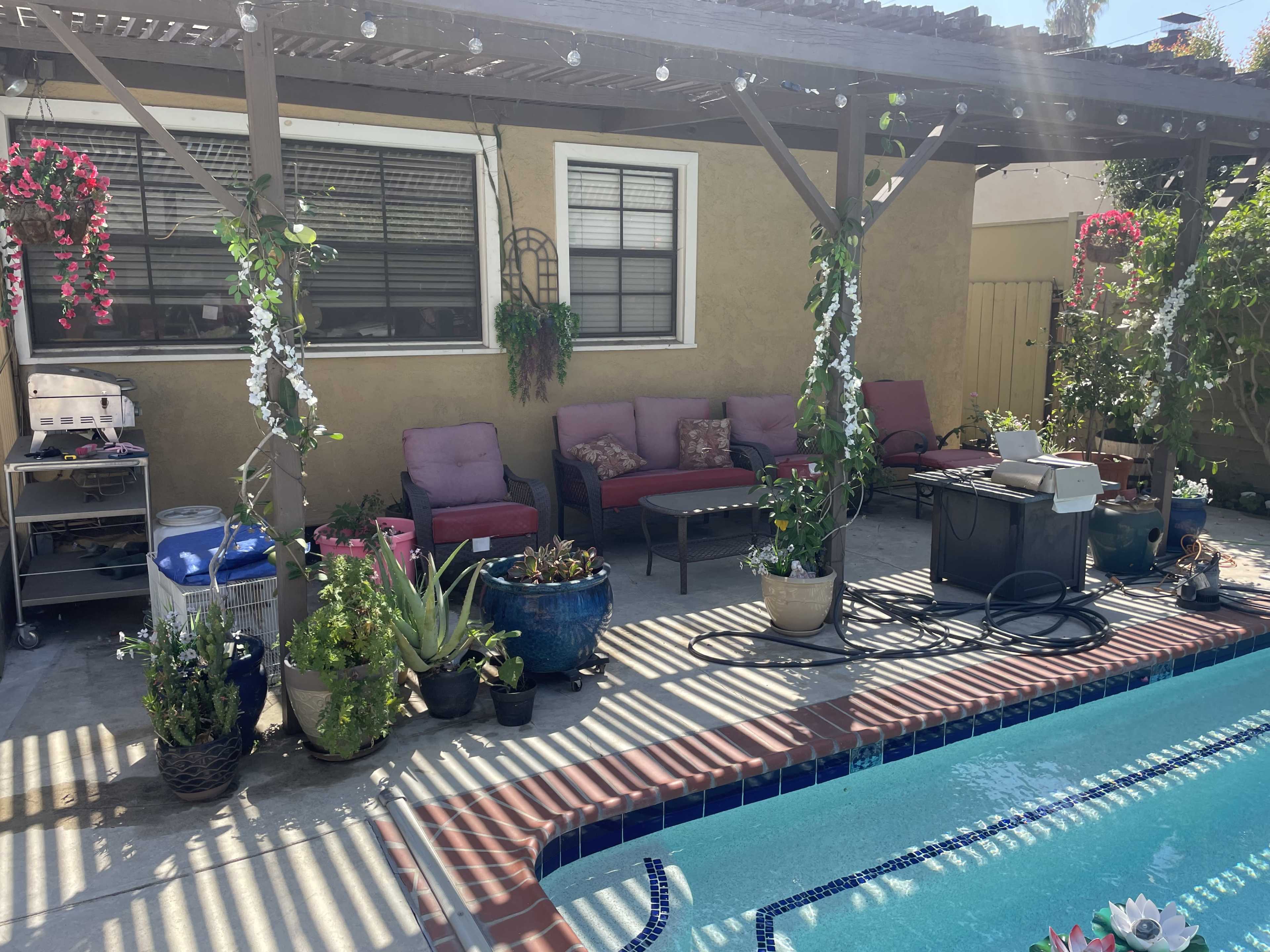 The scene features a backyard patio with a swimming pool, adorned with potted plants and seating arranged under a wooden pergola.