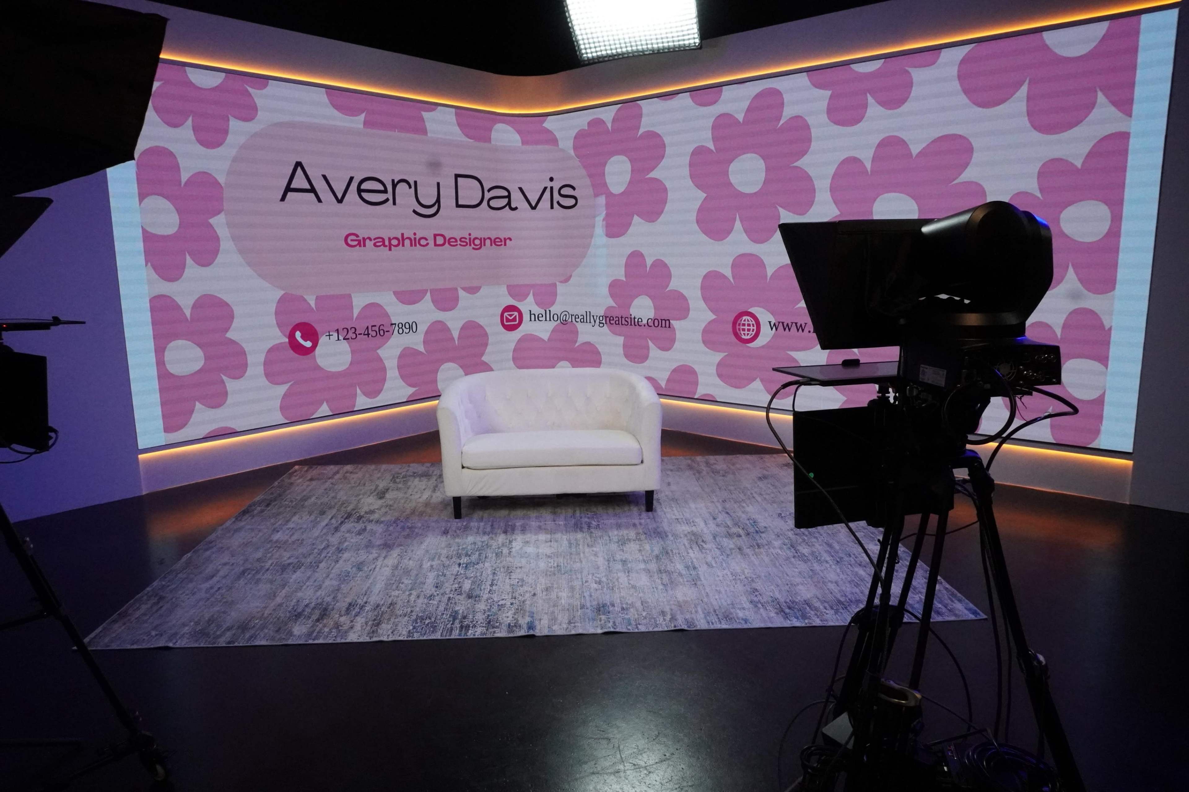 The image shows a television studio set featuring a white couch in front of a vibrant backdrop with pink flowers and the name "Avery Davis" displayed as a graphic designer.