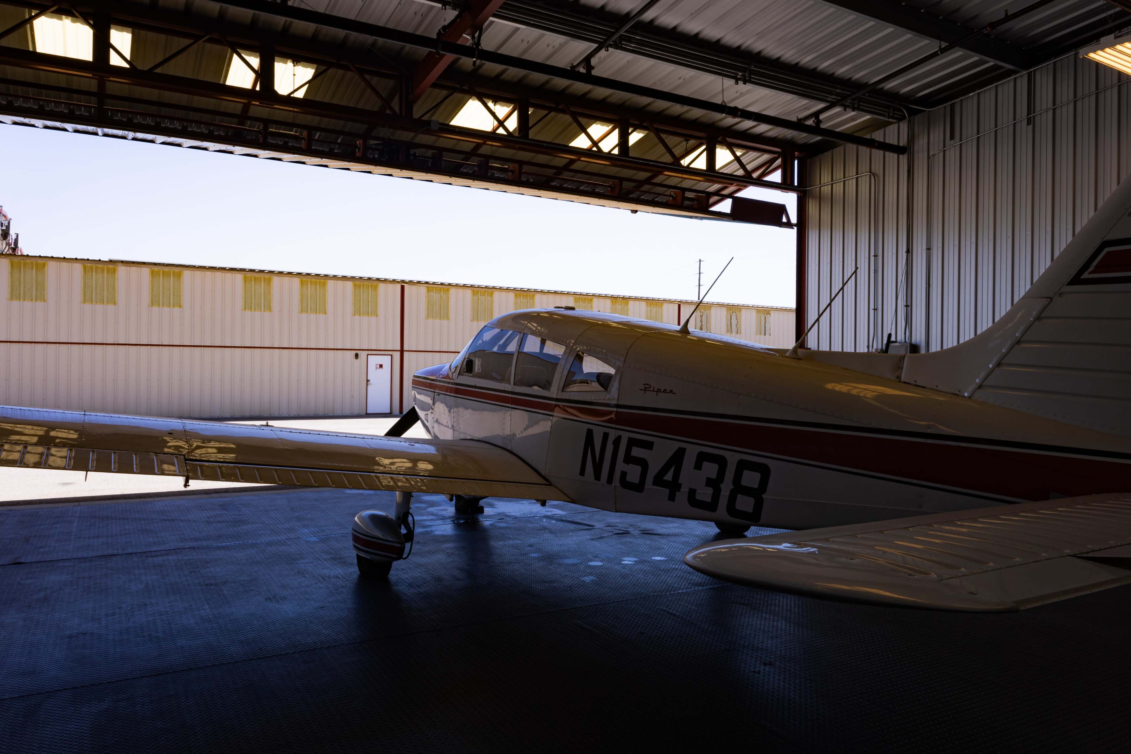 A single-engine airplane is parked inside an aircraft hangar with the hangar door open, revealing a bright, clear sky outside.