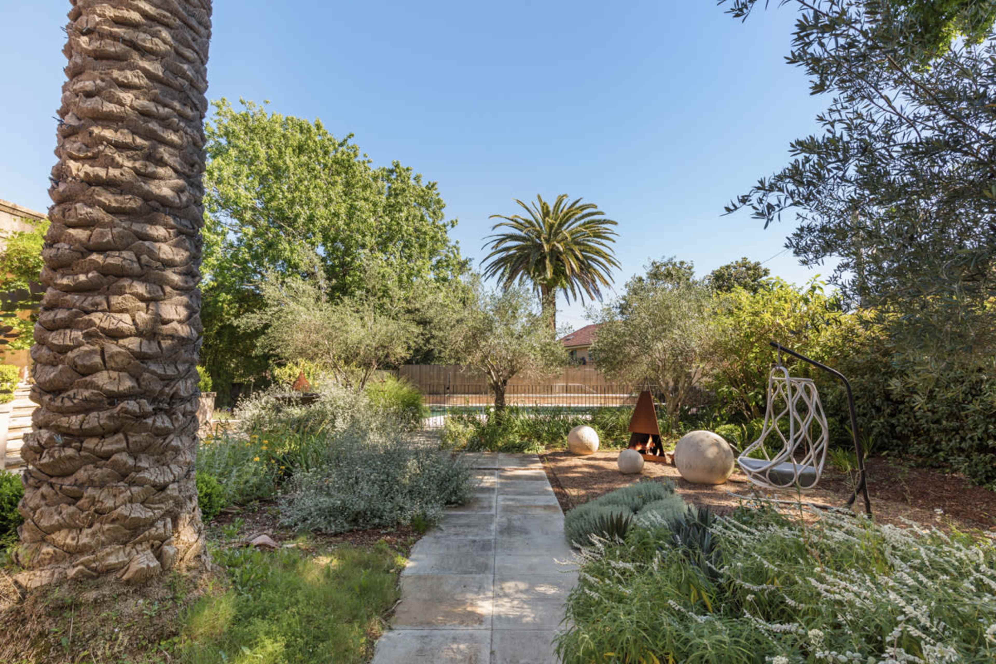 The scene features a landscaped garden with a pathway leading to a pool area, surrounded by various trees, plants, and modern outdoor furnishings.