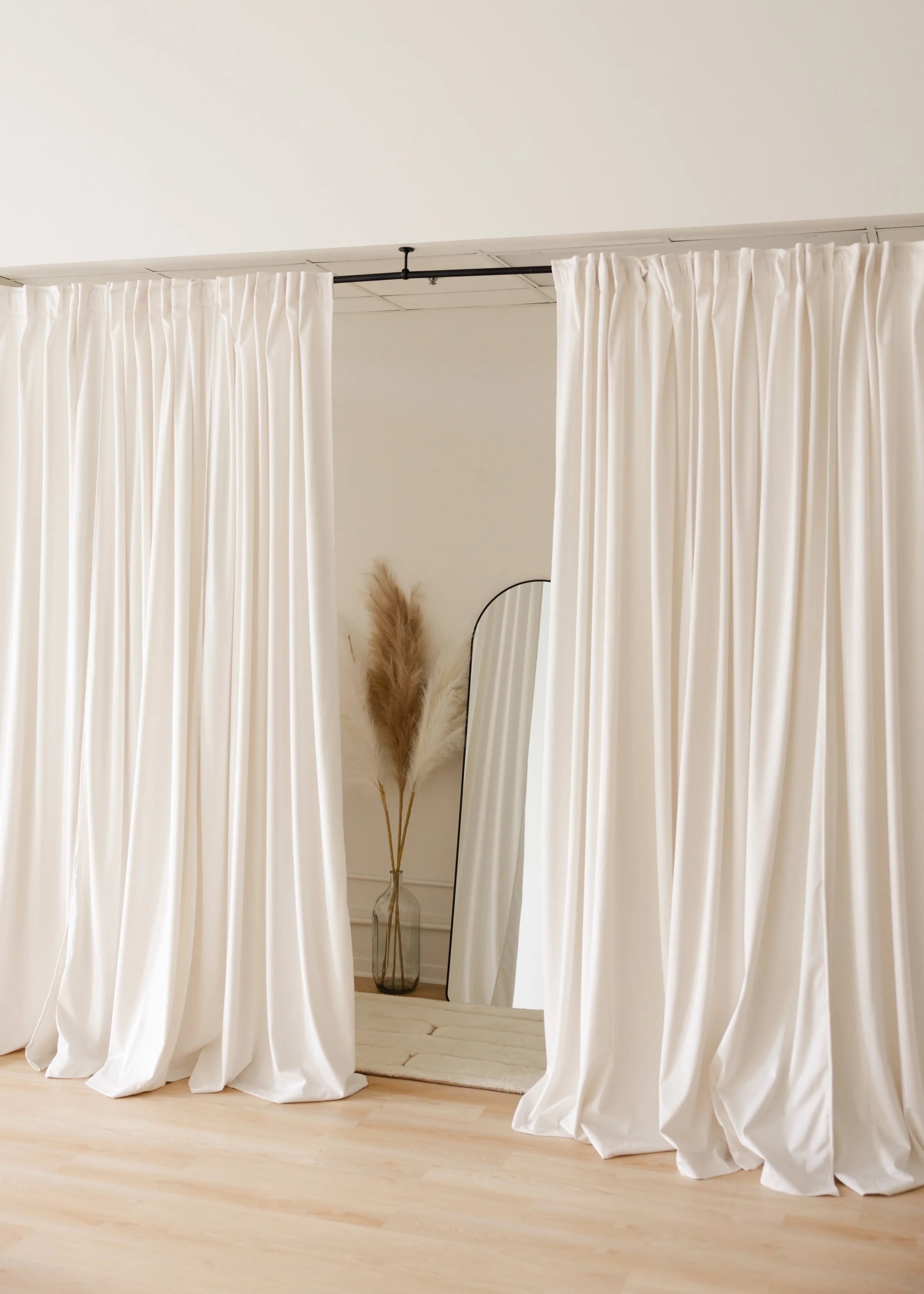 The image shows a pair of white curtains draped over a doorway, revealing a decorative mirror and a vase with dried pampas grass in a minimalist interior setting.