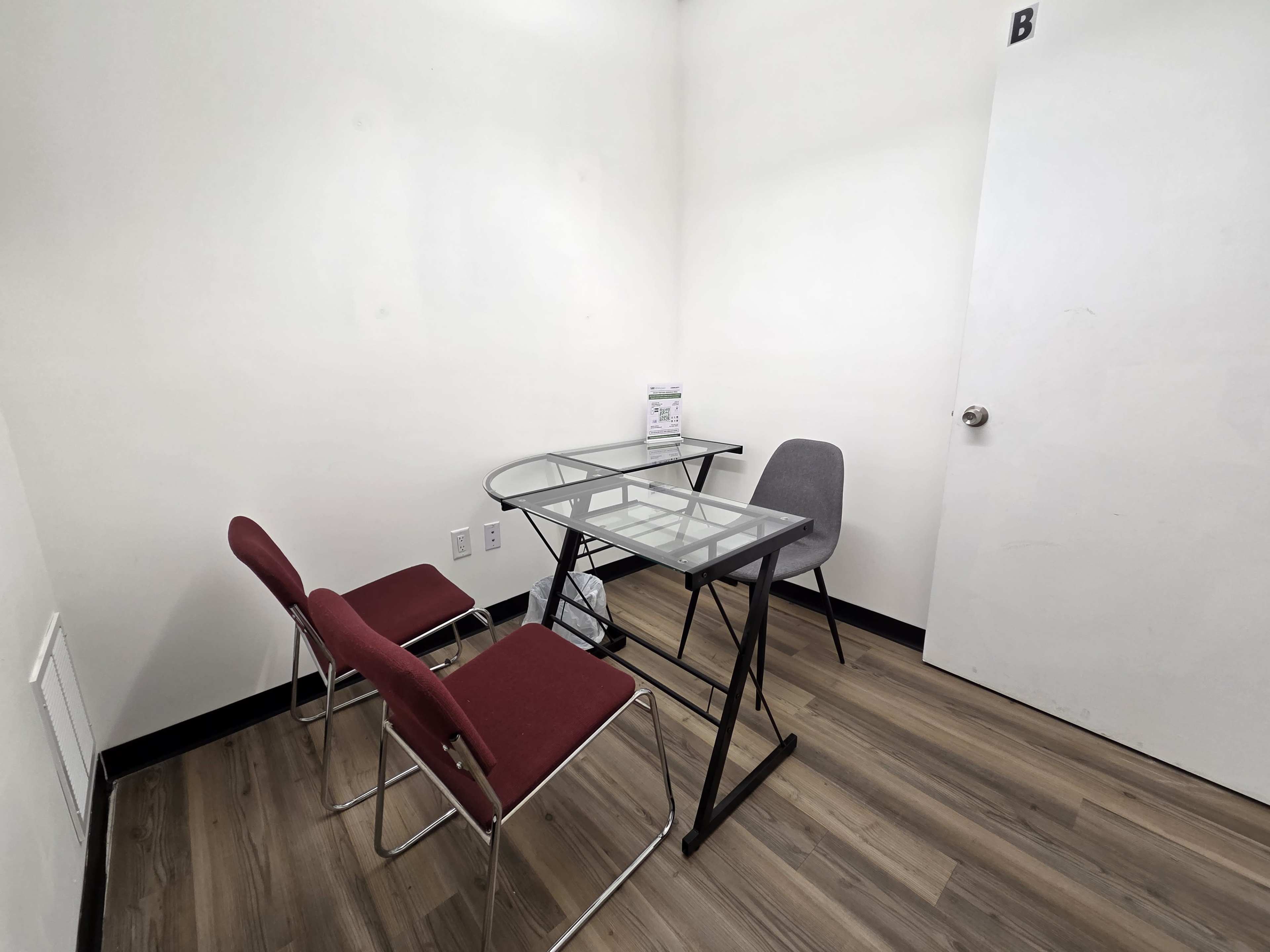 The image shows a small, empty room with a glass-top table and two red chairs, along with a gray chair and a door on one wall.