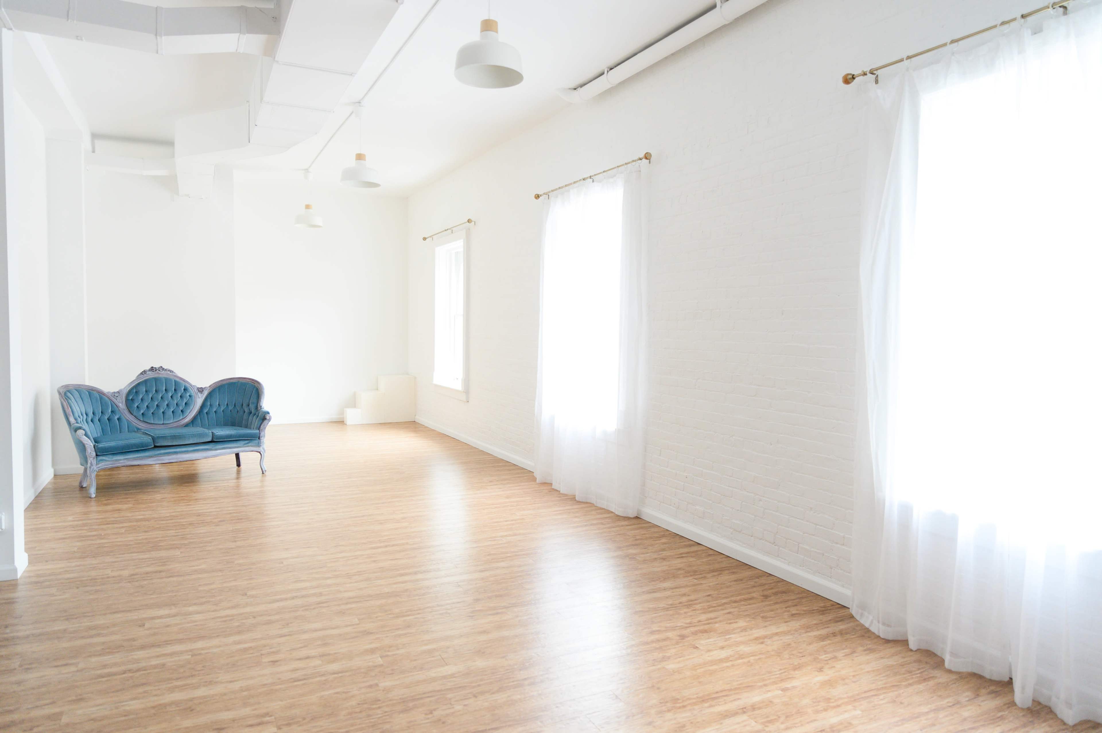 A bright, empty room features light wood flooring, white walls, two large windows with sheer curtains, and a blue velvet sofa against one wall.