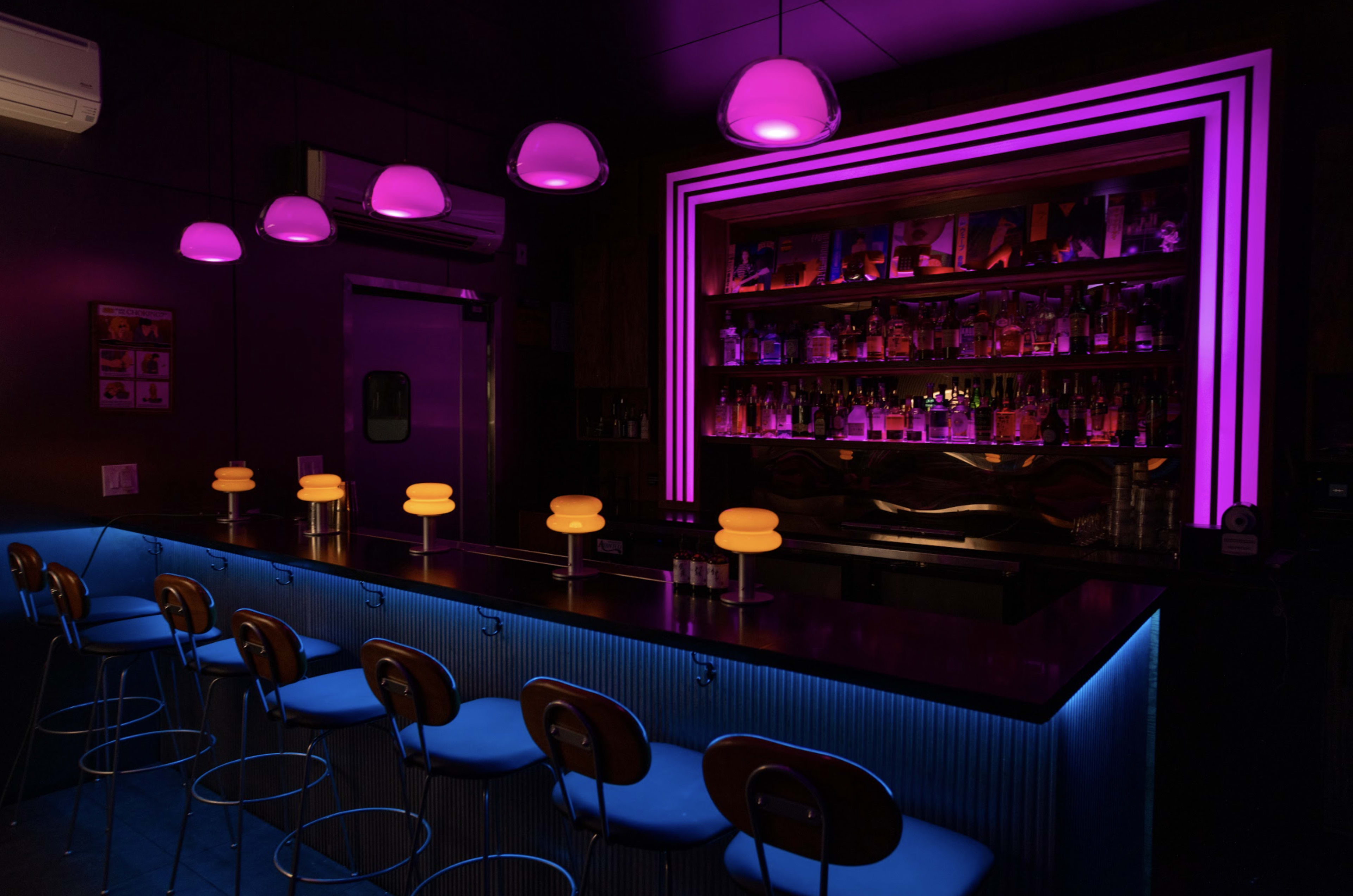 The image shows a sleek bar area with a neon-lit counter and circular stools, illuminated by pink and orange pendant lights.