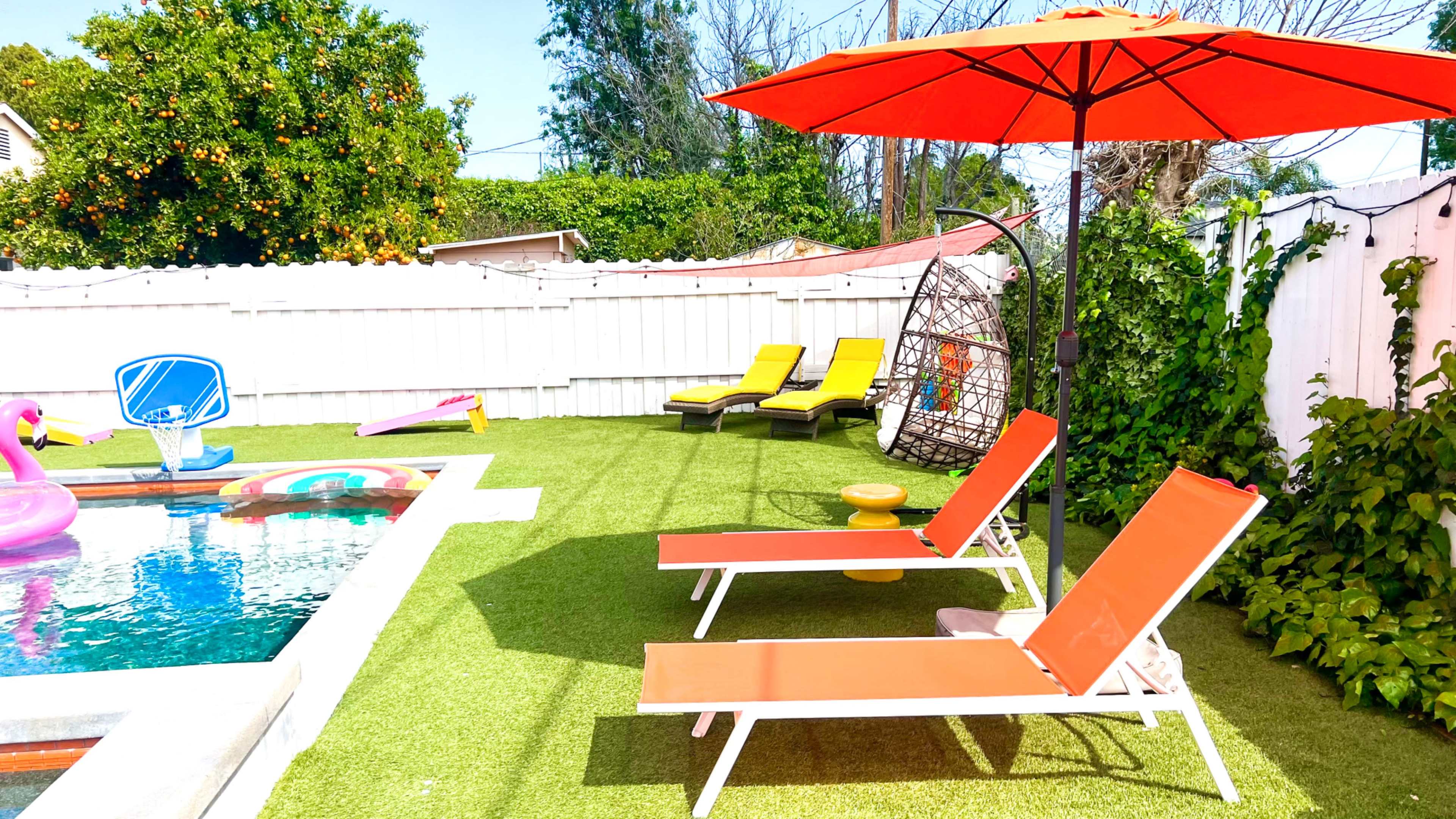 The image shows a backyard pool area with orange lounge chairs, an umbrella, and a hanging chair, surrounded by green grass and a white fence.