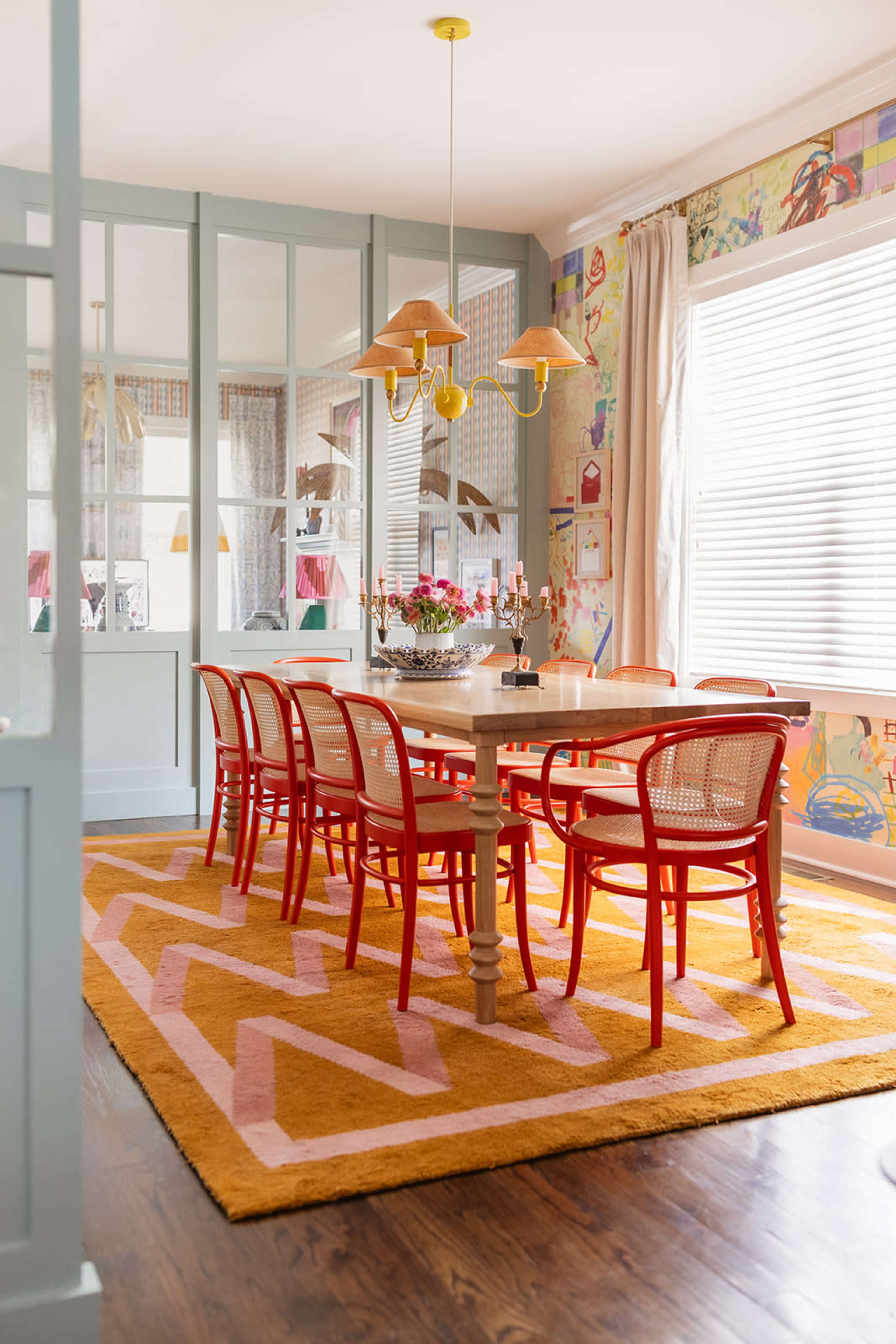 A bright dining room features a round table surrounded by red chairs, with a yellow rug and patterned walls.