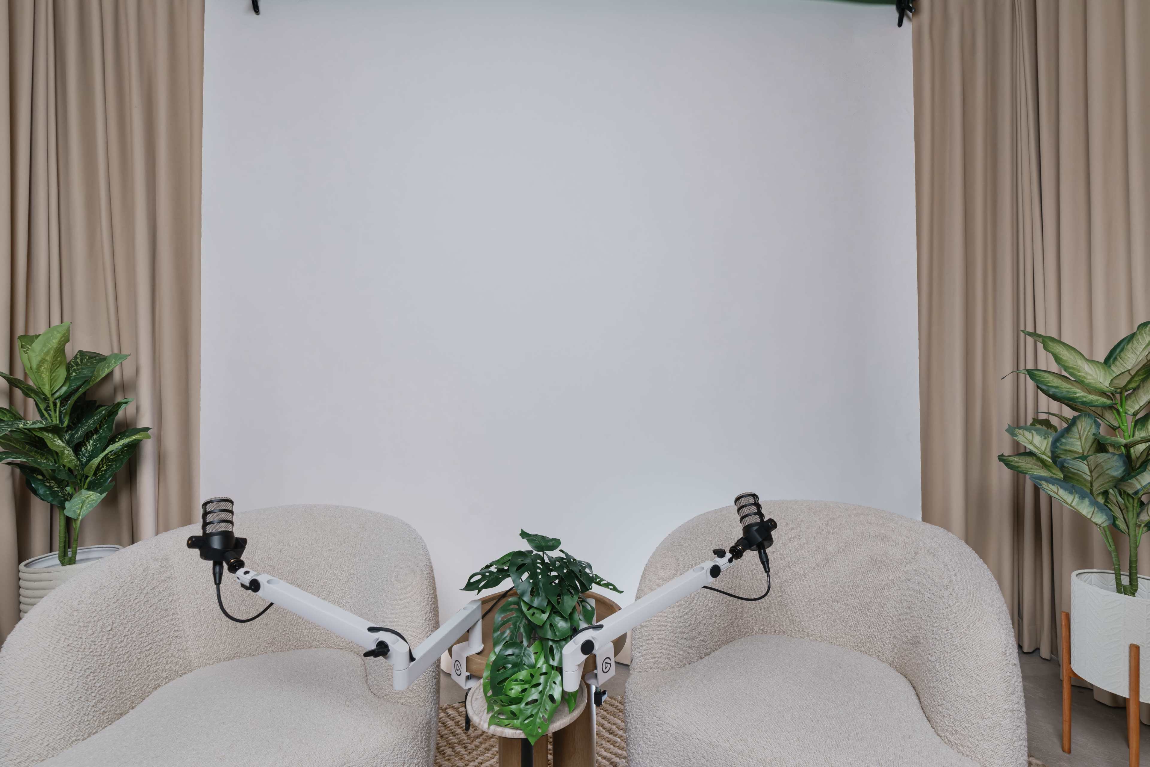 Two light-colored chairs with white adjustable arms and a potted plant between them in front of a blank backdrop and curtain.
