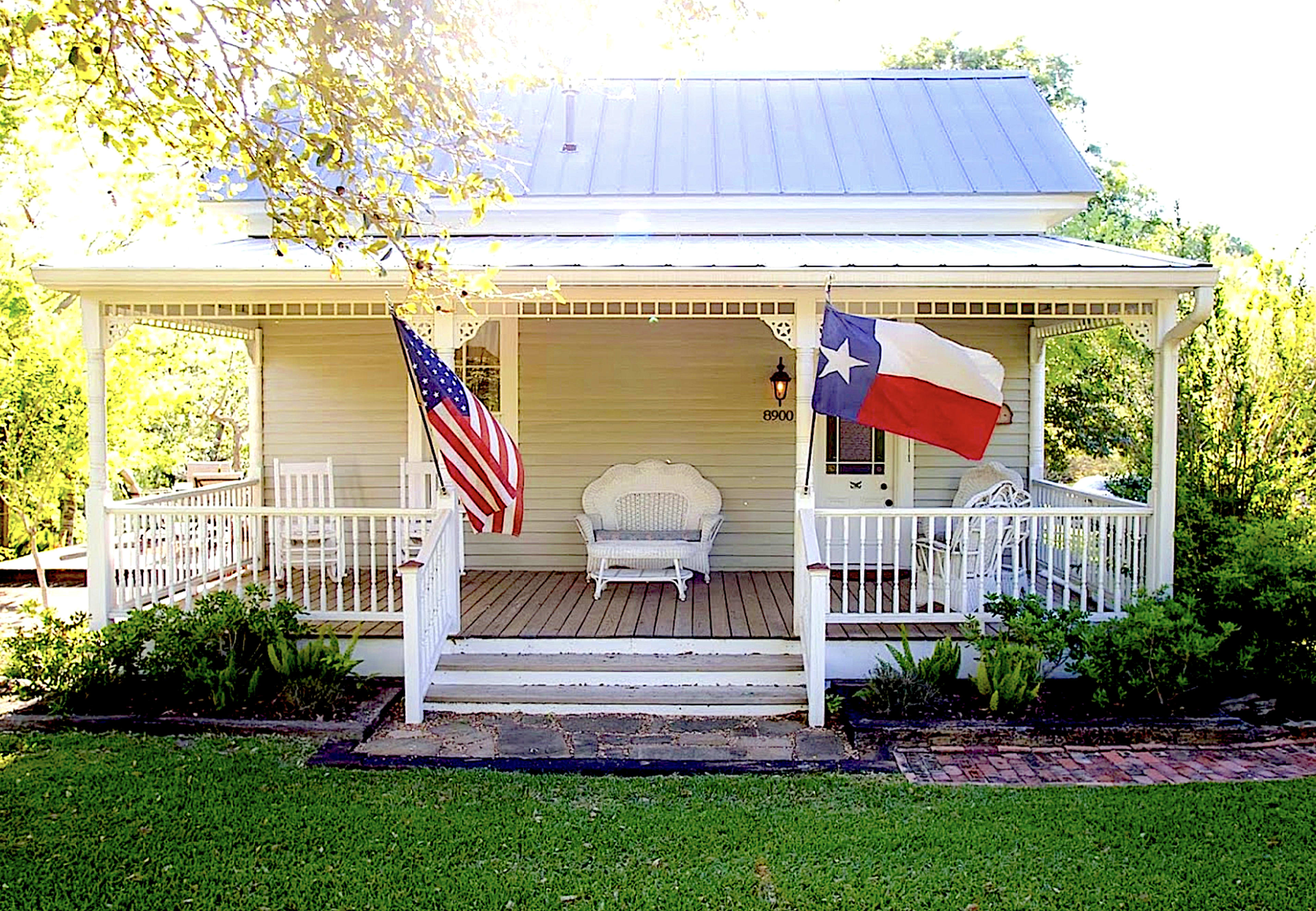 The image shows a traditional house with a front porch, featuring an American flag and a Texas flag displayed on opposite sides.