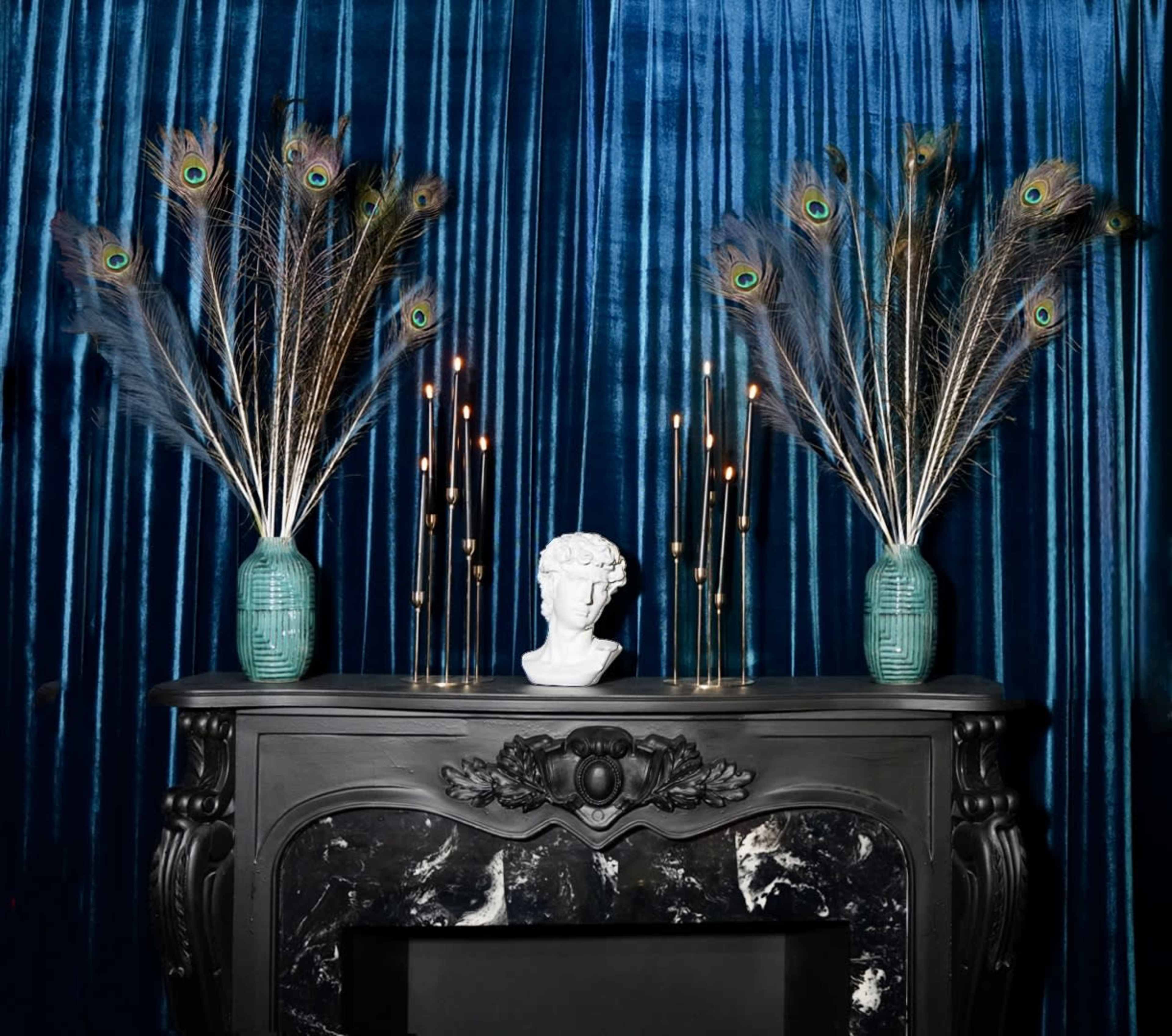 A black fireplace mantel displays a white bust, flanked by vases with peacock feathers and lit candles against a blue velvet backdrop.