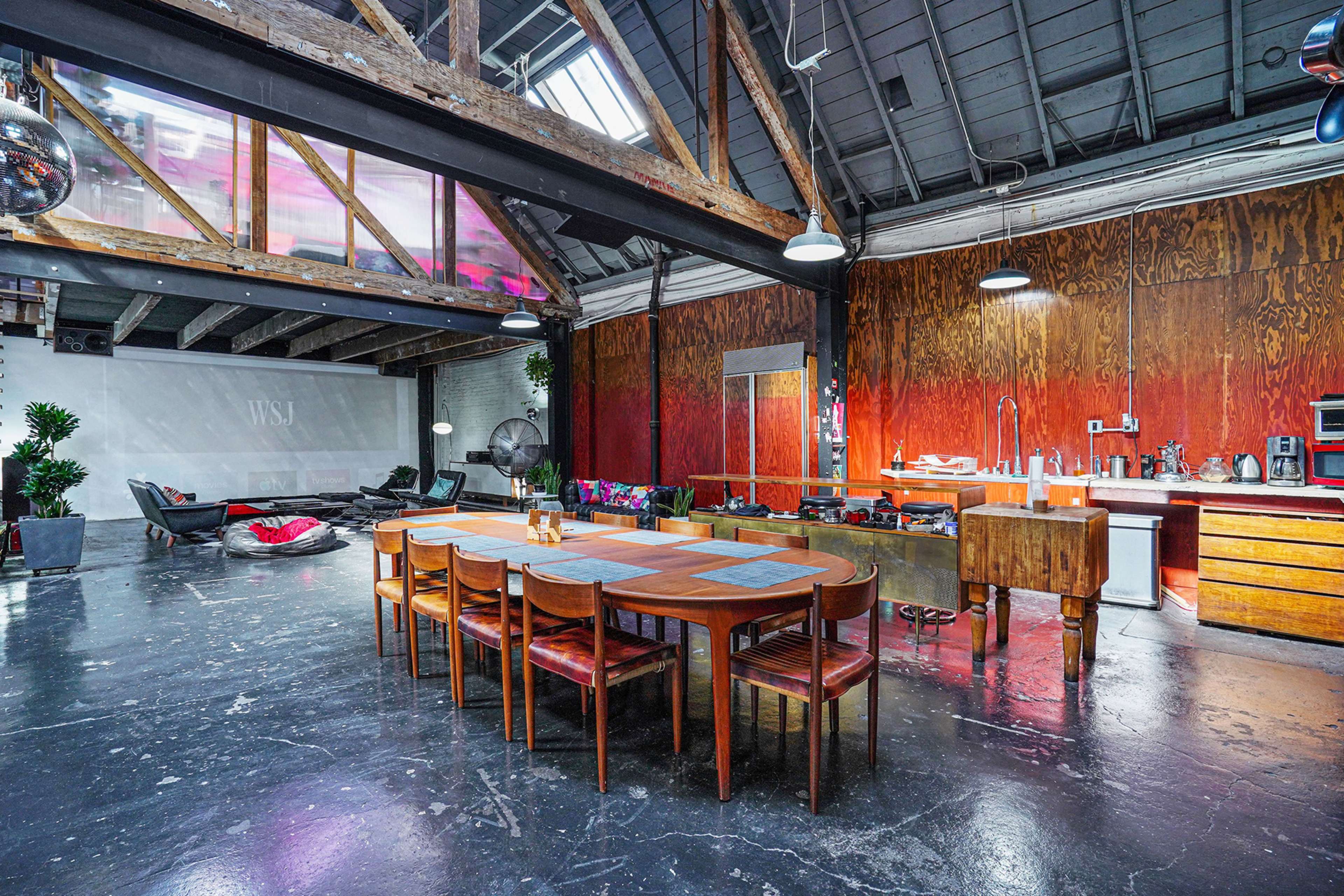 A spacious industrial-style dining area features a long wooden table surrounded by chairs, with a kitchen area and large windows allowing light to fill the space.