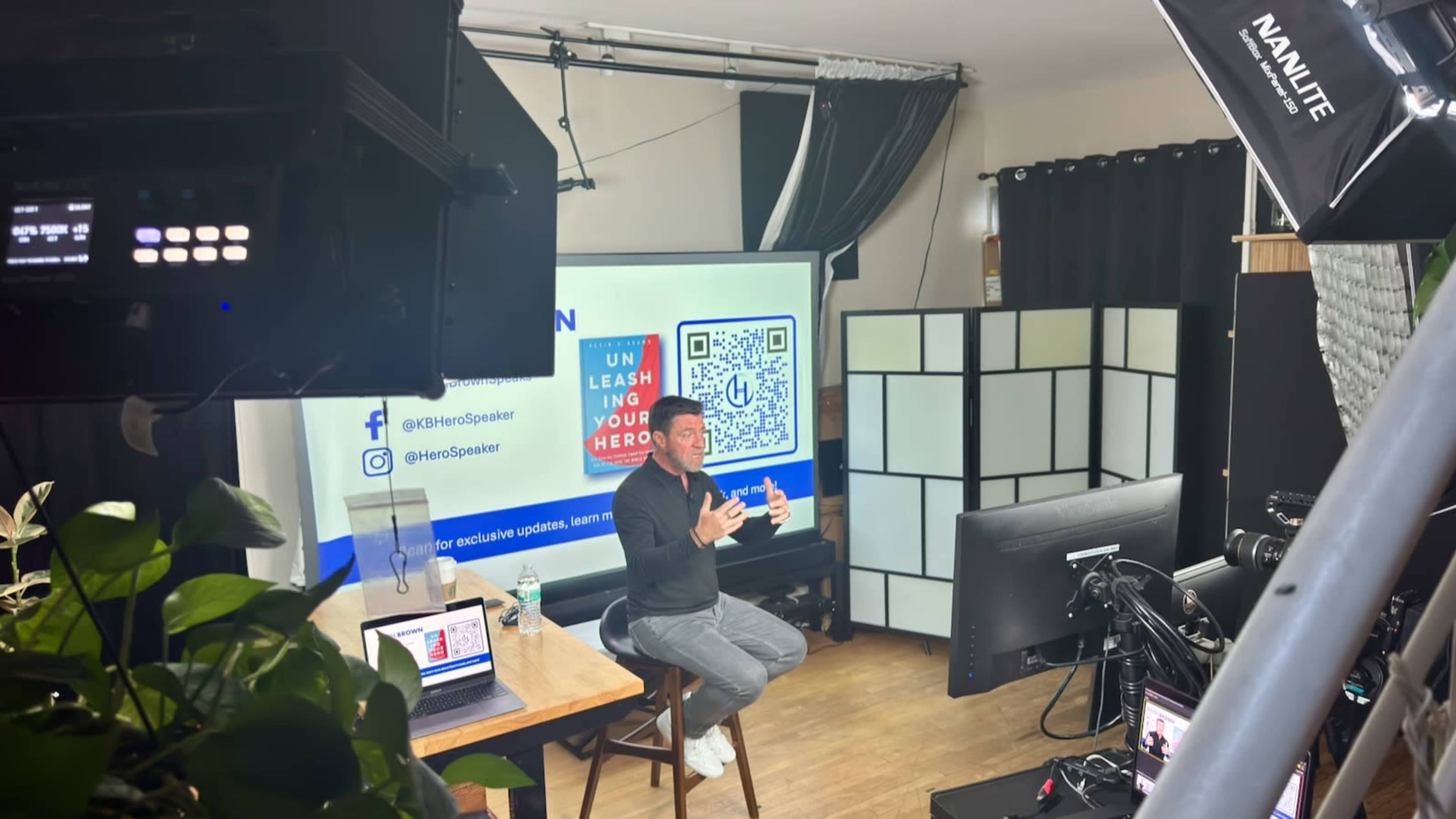 A speaker presents in front of a large screen in a well-lit room equipped with cameras and studio lighting.