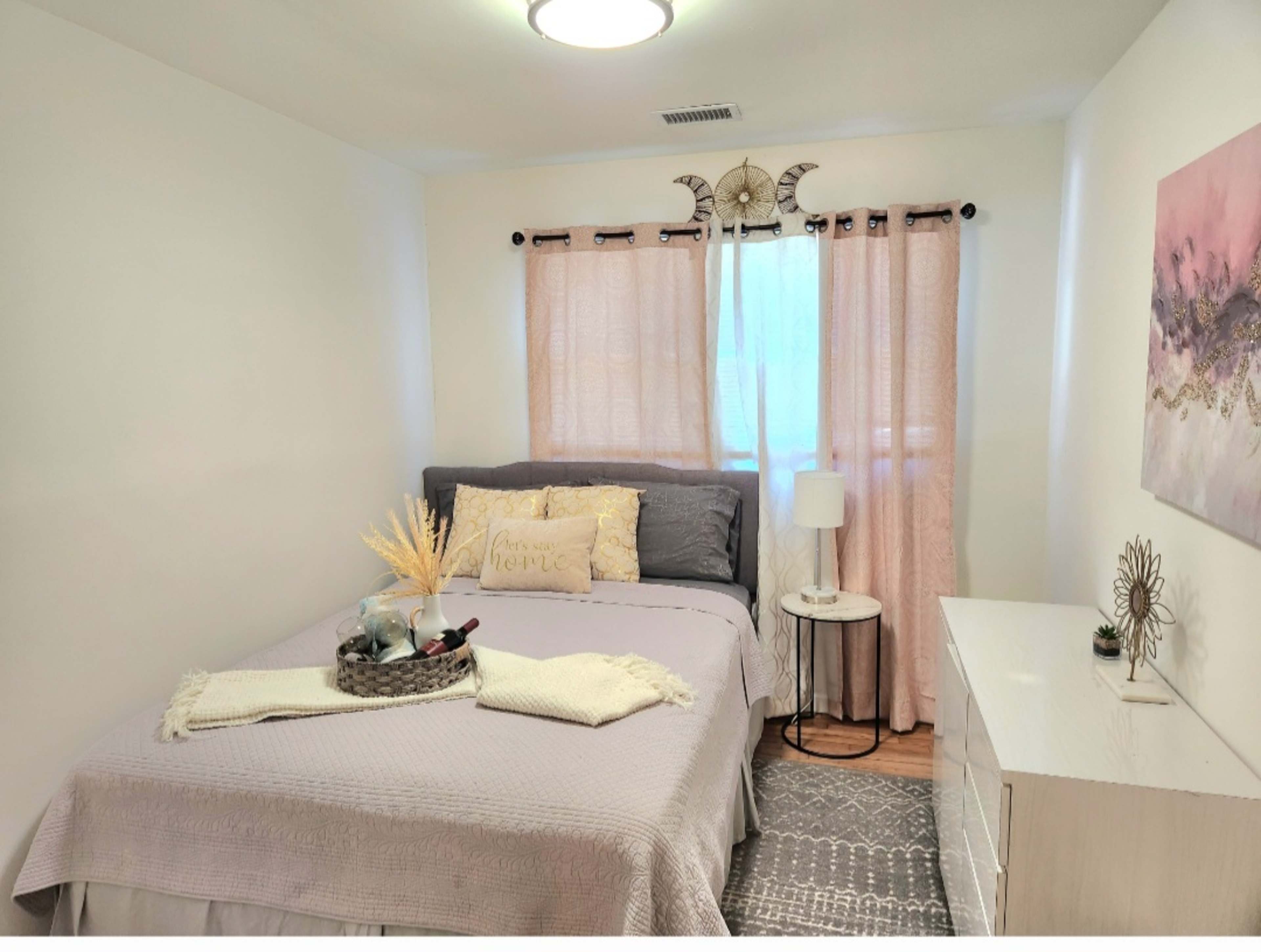 A well-lit bedroom featuring a bed with gray bedding, a bedside lamp, and a dresser against a white wall.