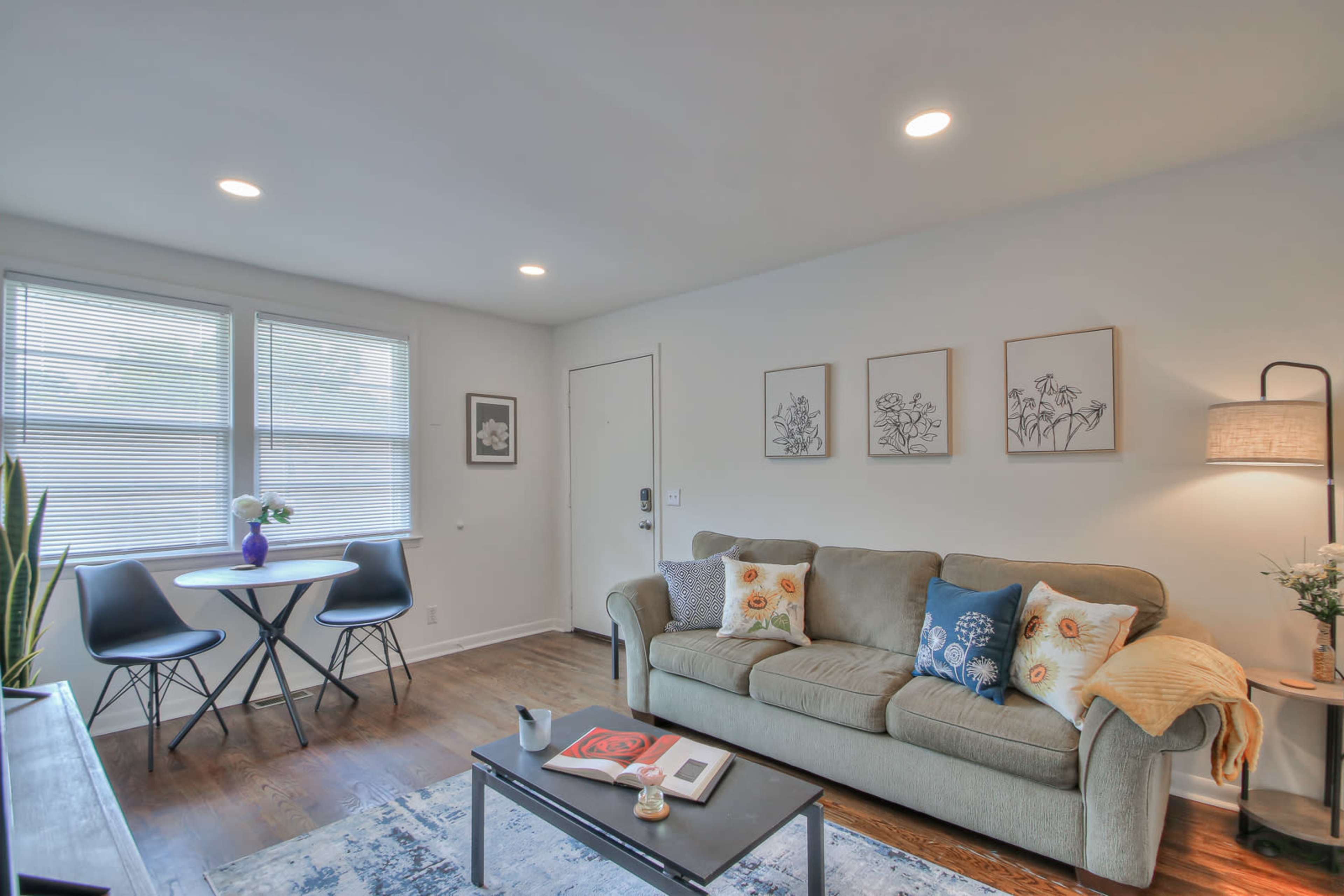 The image shows a cozy living room with a sofa, a small dining table, and framed artwork on the walls.