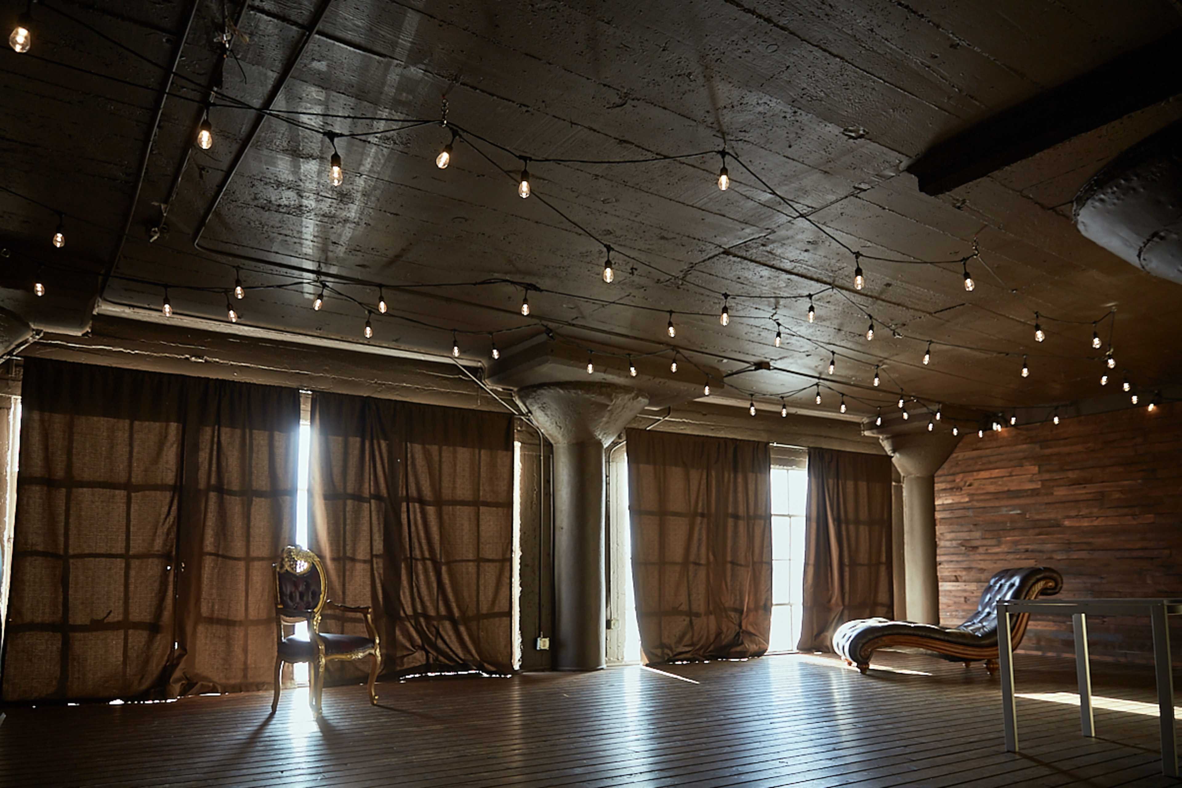 A spacious room with wooden flooring, large brown curtains, and string lights hanging from the ceiling.