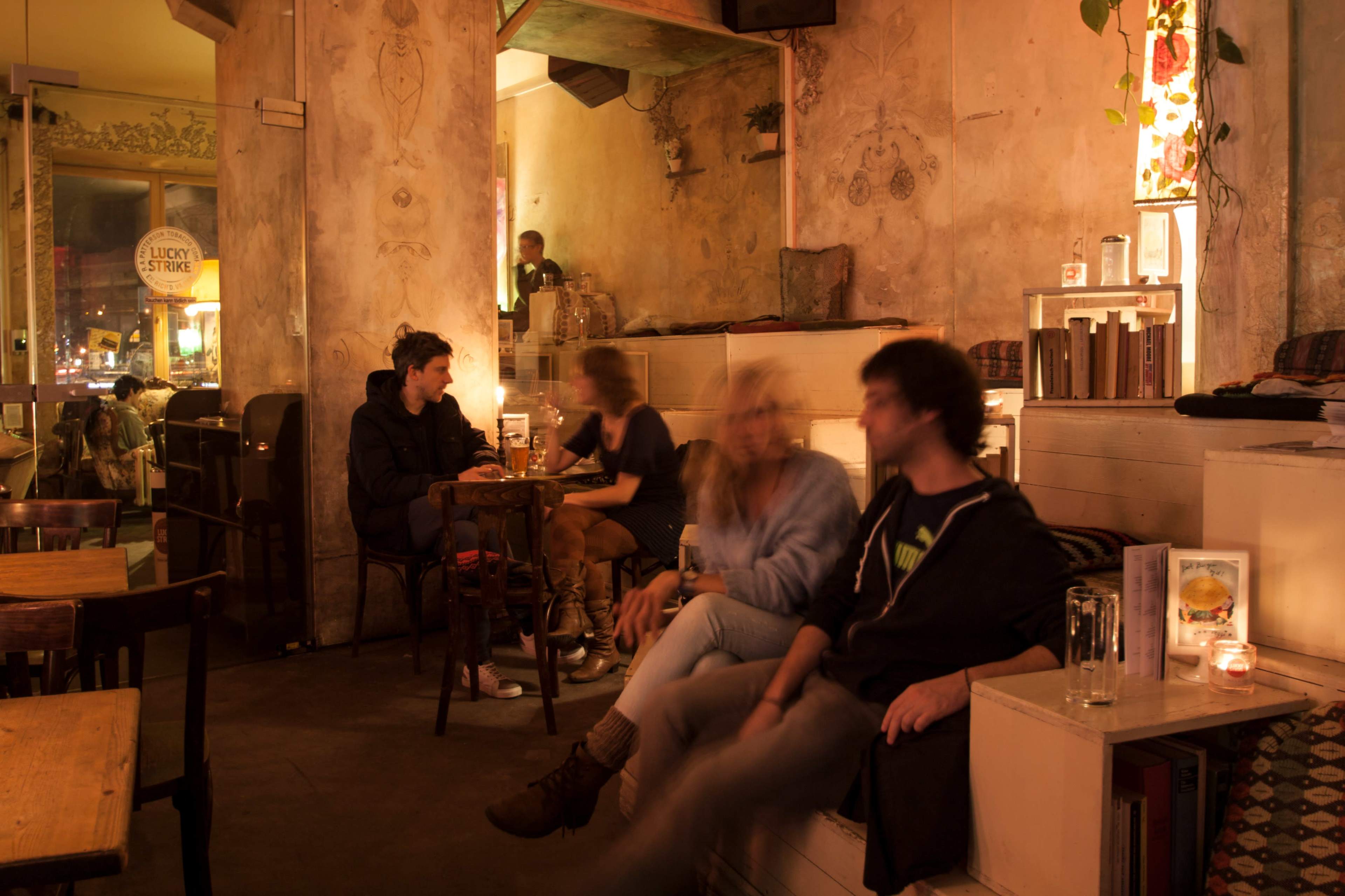 A cozy café interior with soft lighting, where several people sit at tables while others lounge on a low platform.