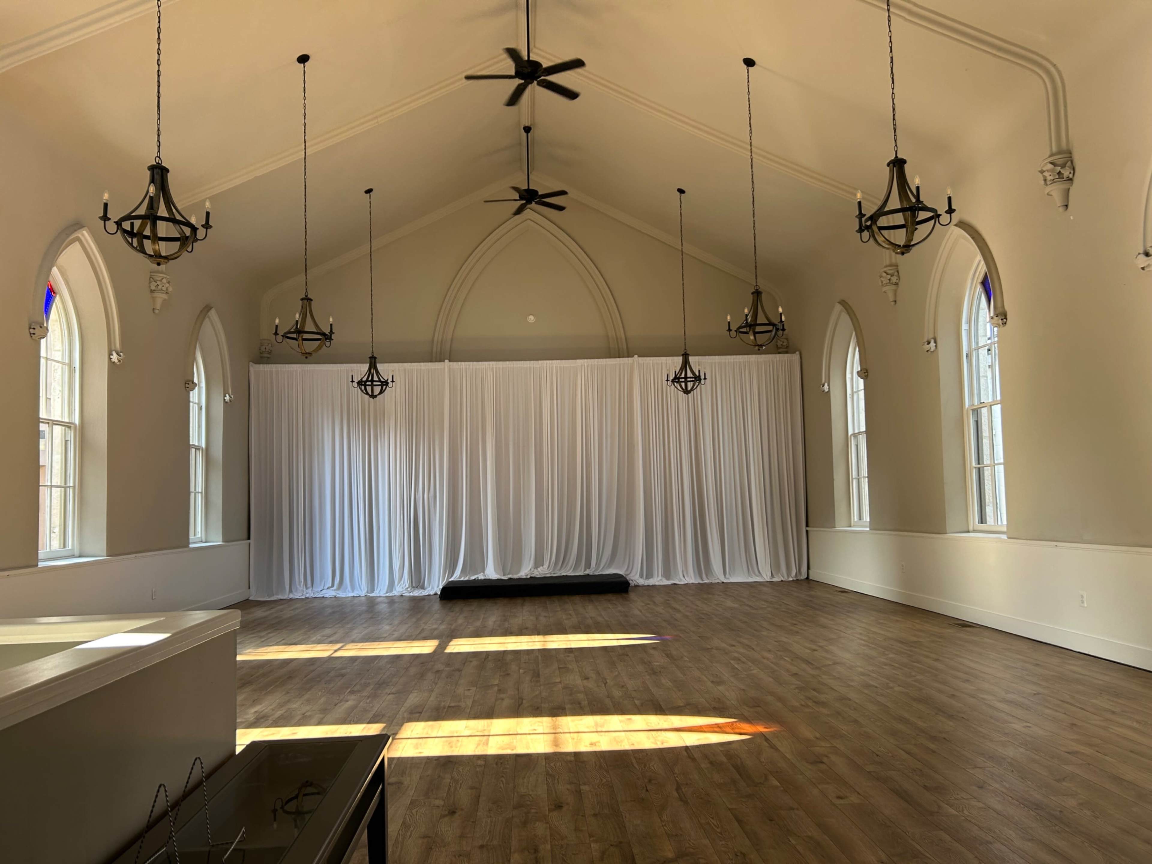 The image shows a spacious, empty hall with high ceilings, large windows, and white drapery at one end.