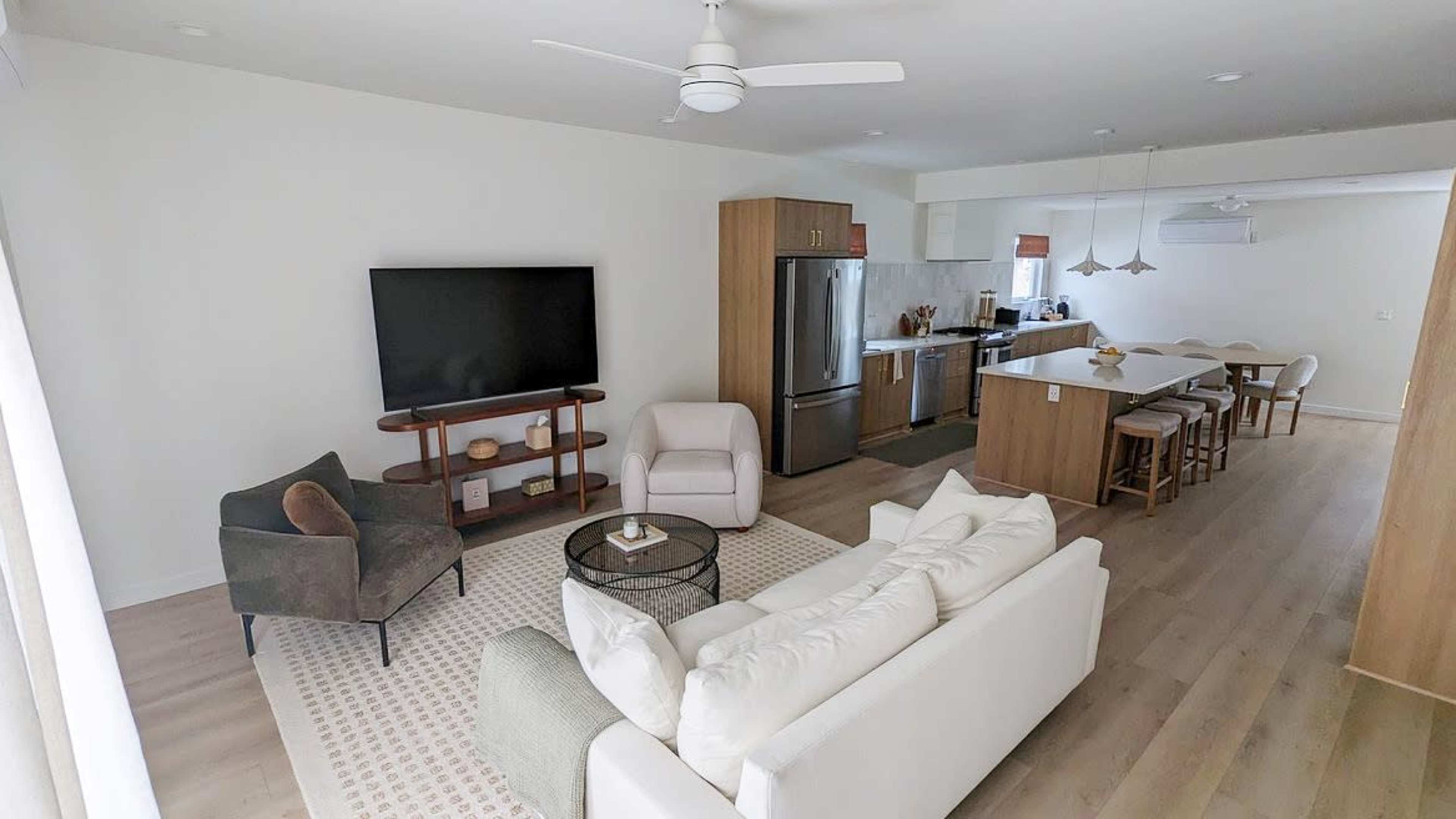 The image shows a modern living room and kitchen area featuring a white sofa, a brown armchair, a television, and a kitchen with wooden cabinetry and bar seating.