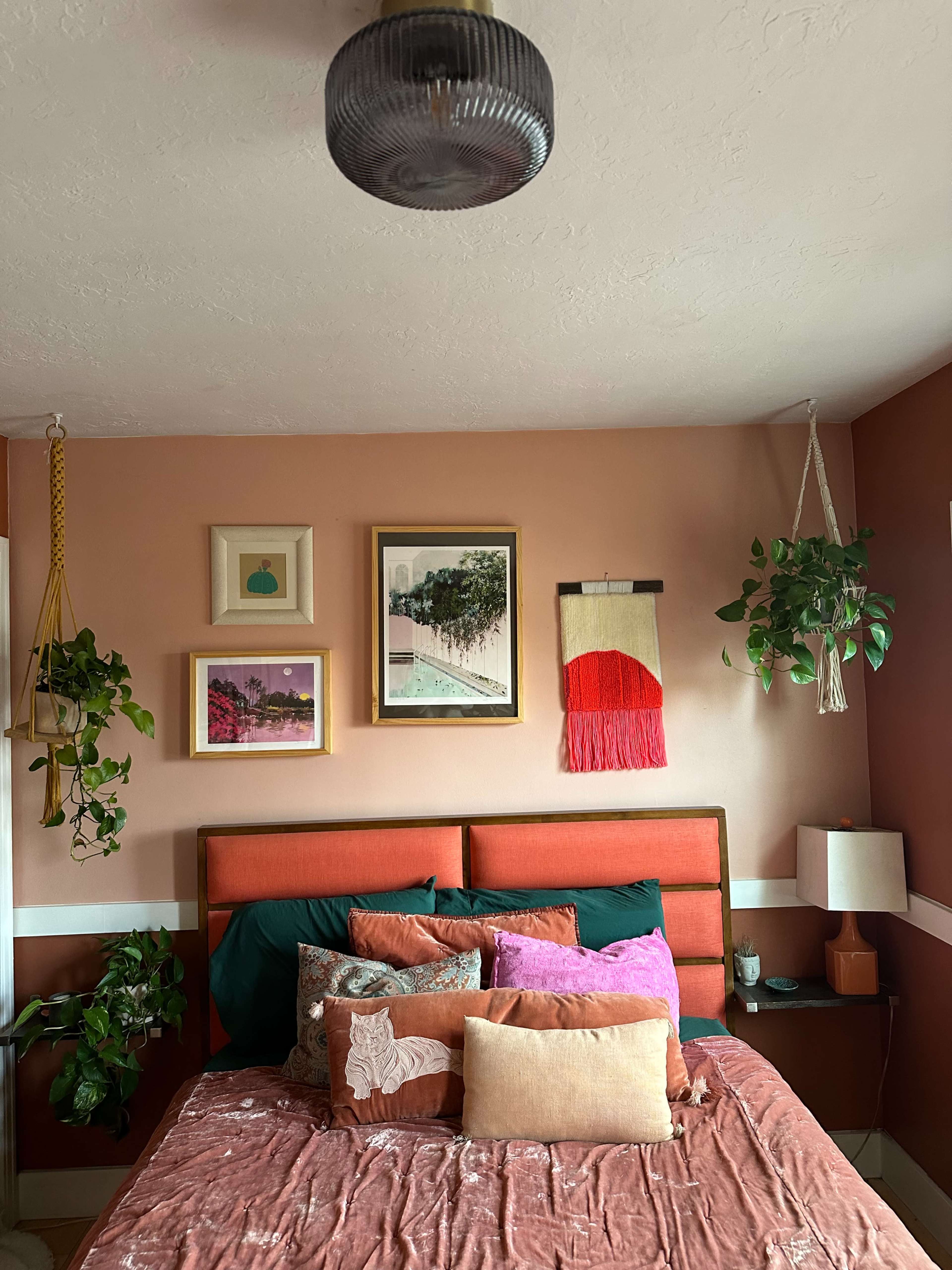 A cozy bedroom with a bed adorned with colorful pillows, framed artwork on the walls, and several hanging plants.