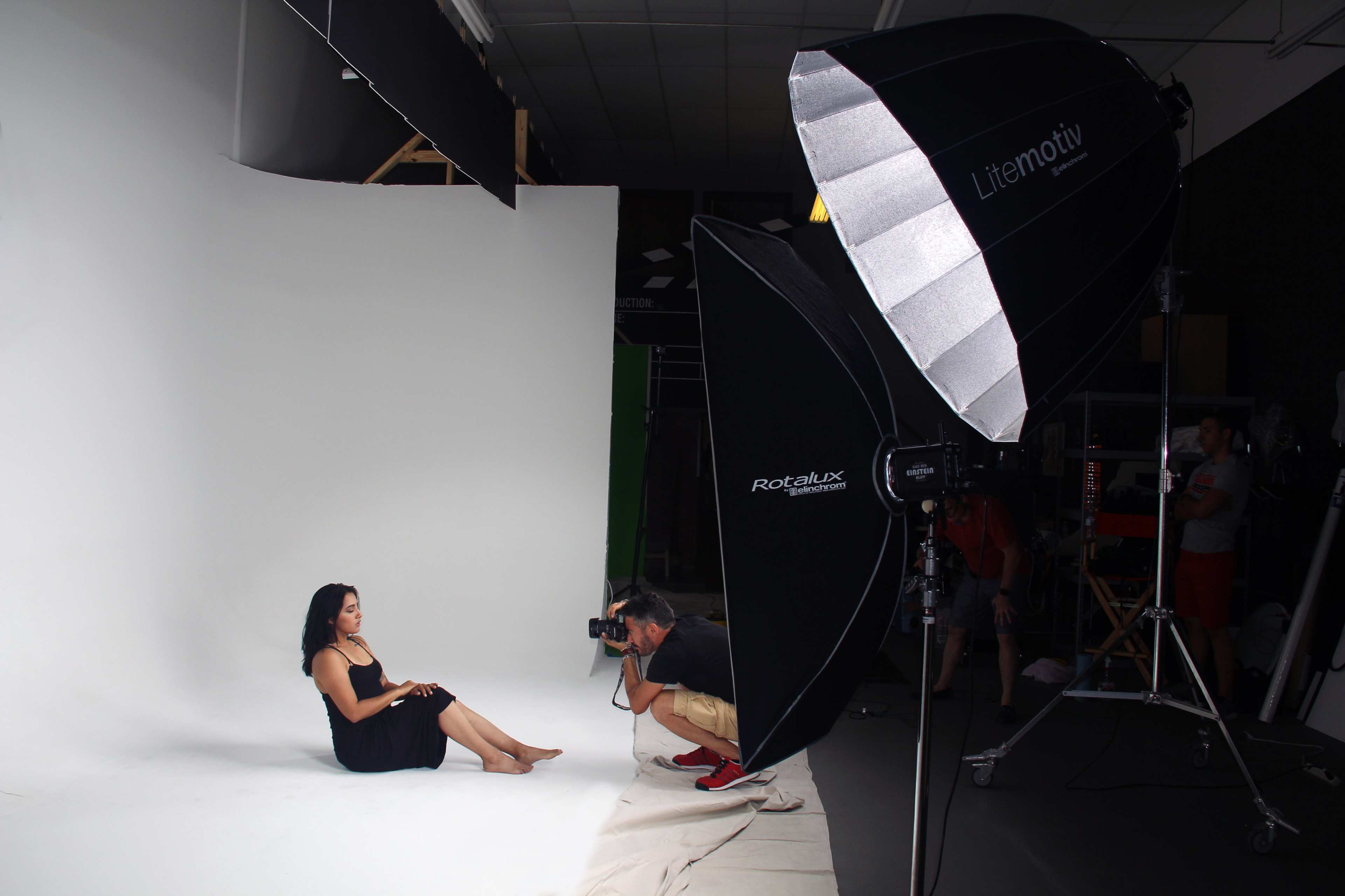 A photographer captures a model posing on a white backdrop in a studio equipped with a large softbox light.