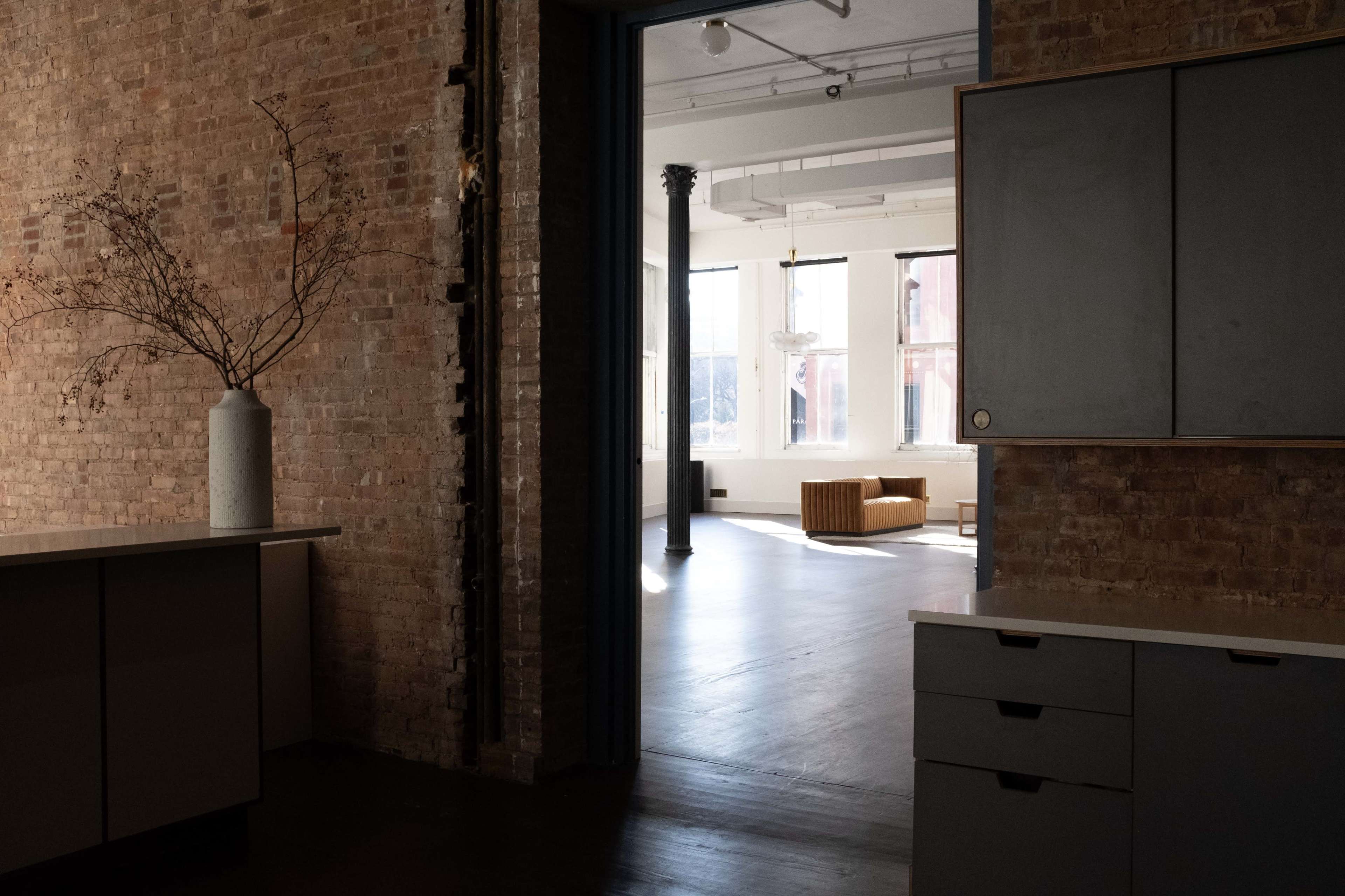 A spacious, minimalist interior features a small kitchen area with a vase and a view into a bright living space with a couch and large windows.