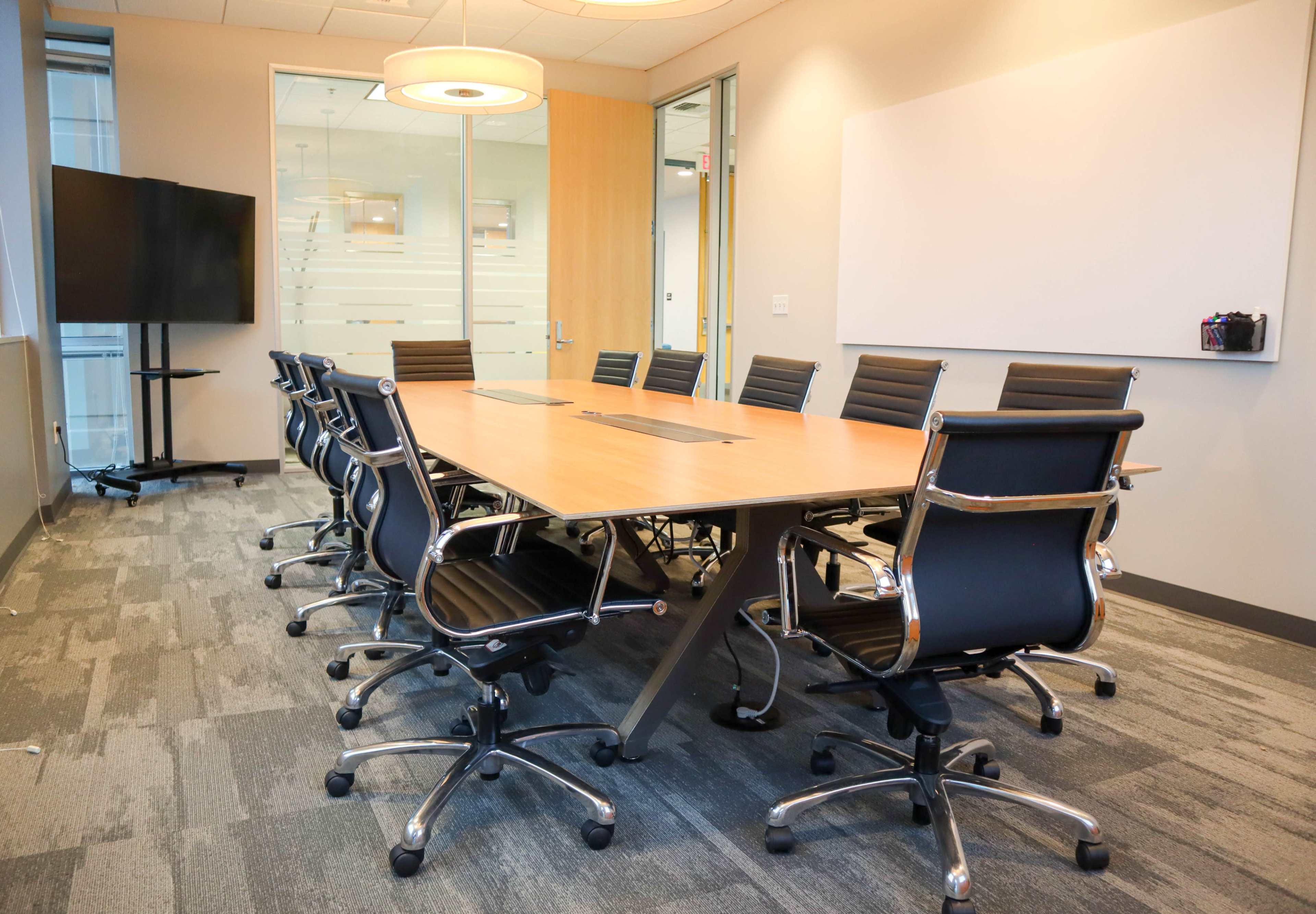 A conference room features a large wooden table surrounded by black rolling chairs, with a flat-screen TV and a whiteboard visible in the background.