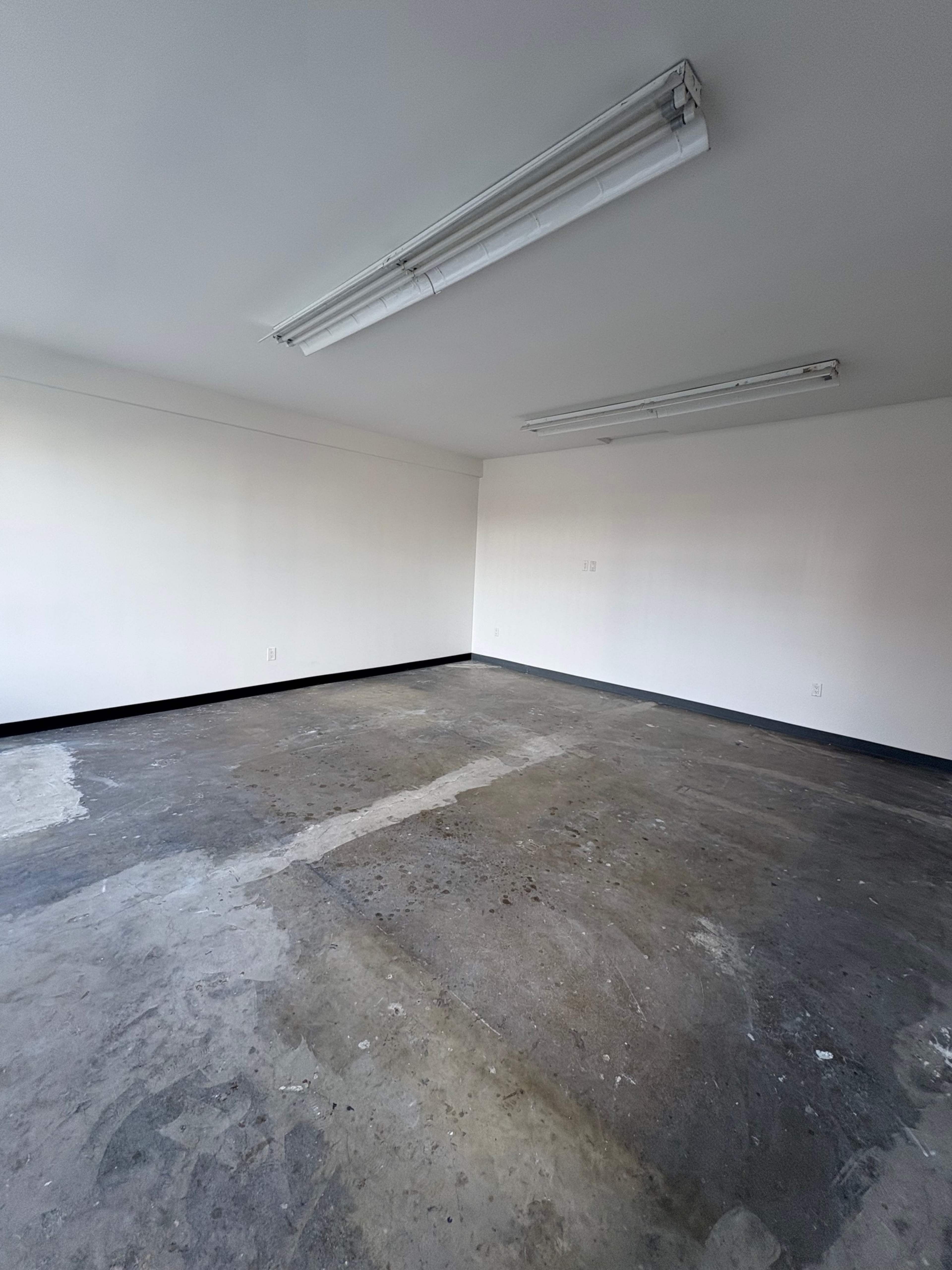 An empty room with a concrete floor, white walls, and fluorescent lighting.