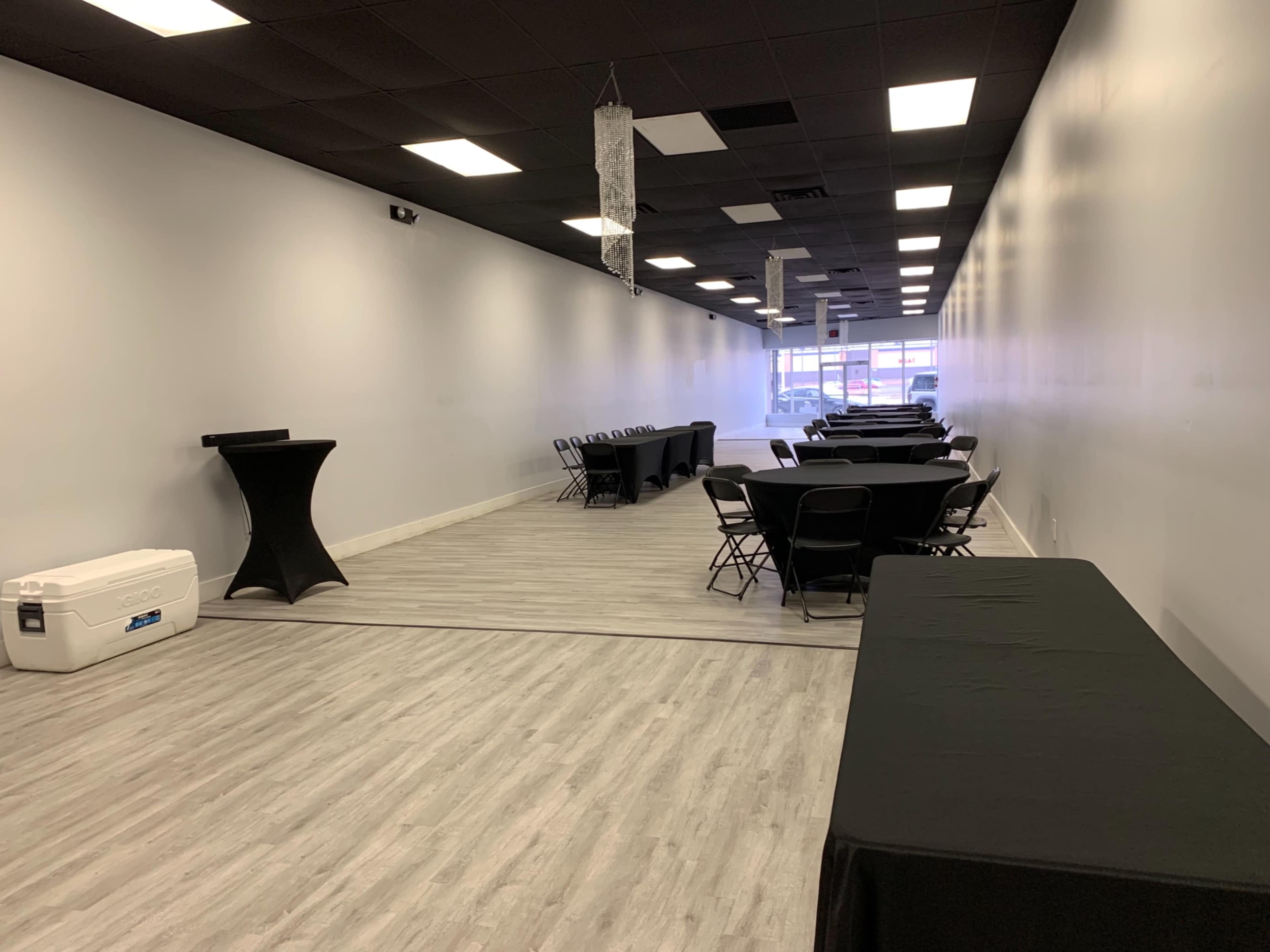 A long, narrow event space with several rows of black tables and chairs arranged in a central area, flanked by a bar-height table and a cooler.