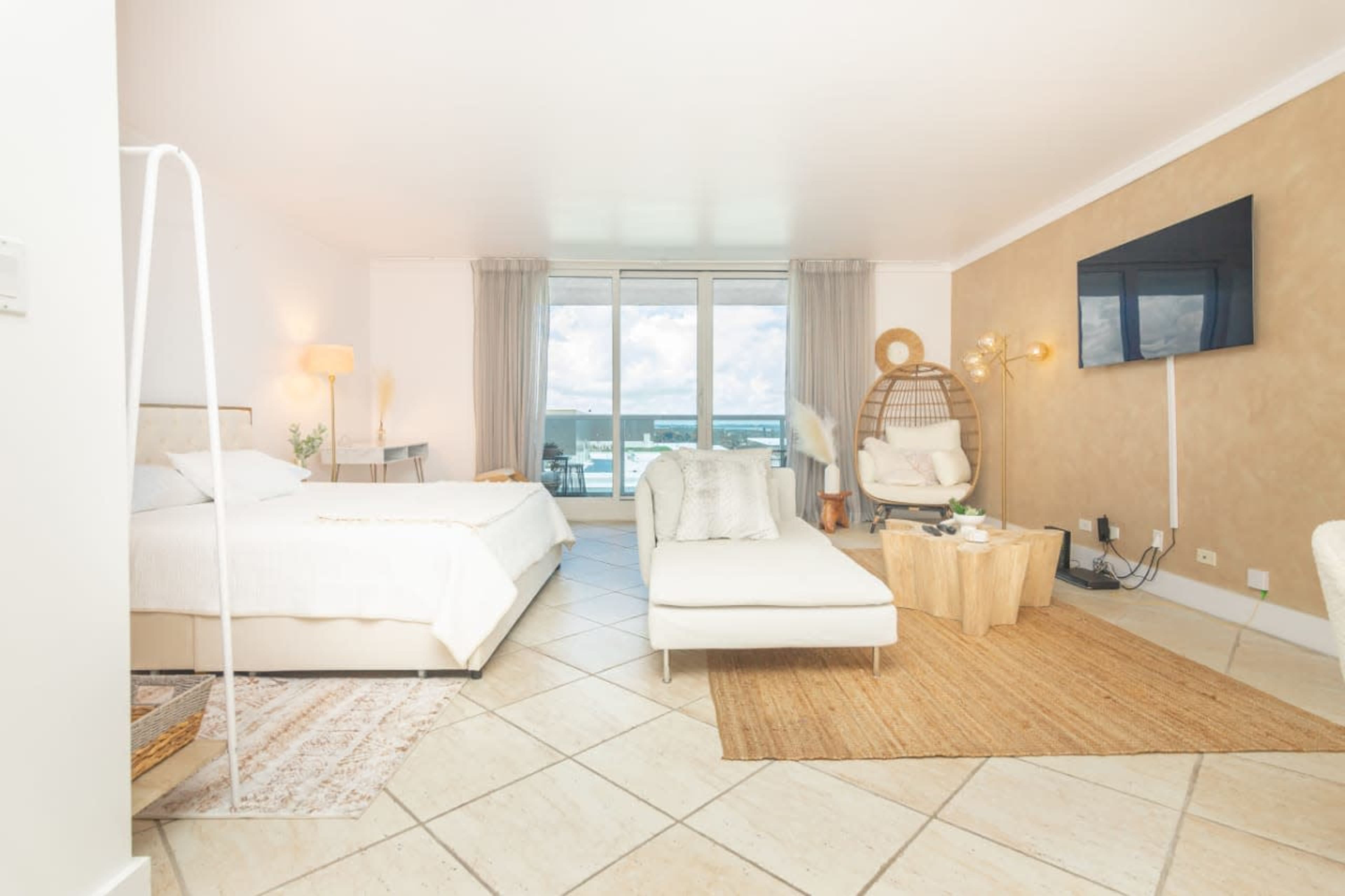 A bright, modern hotel room features a bed, a lounge chair, and a large window with a view of the outside.