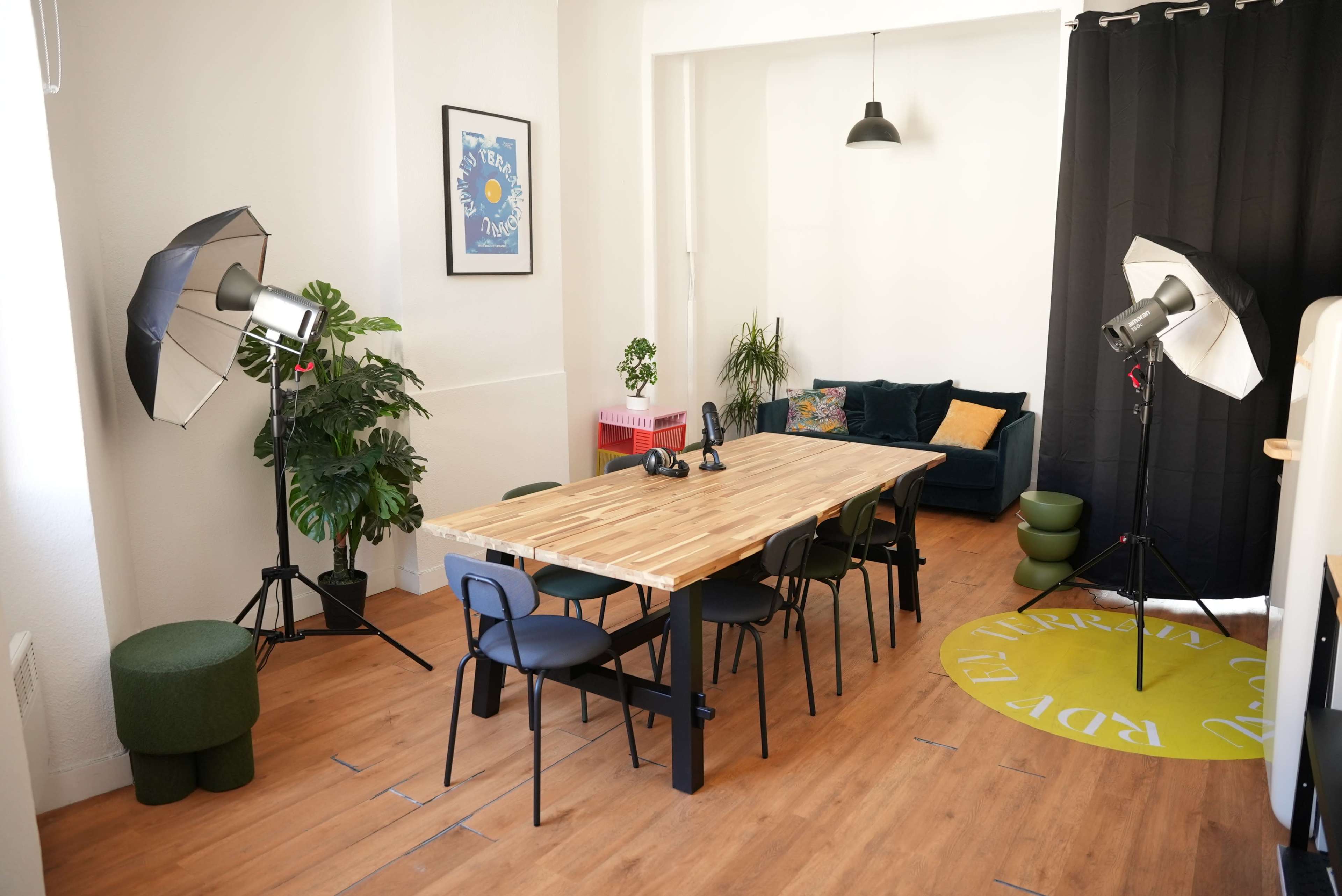 A well-lit room features a large wooden table surrounded by black chairs, with photography lights positioned around and a green plant in the corner.