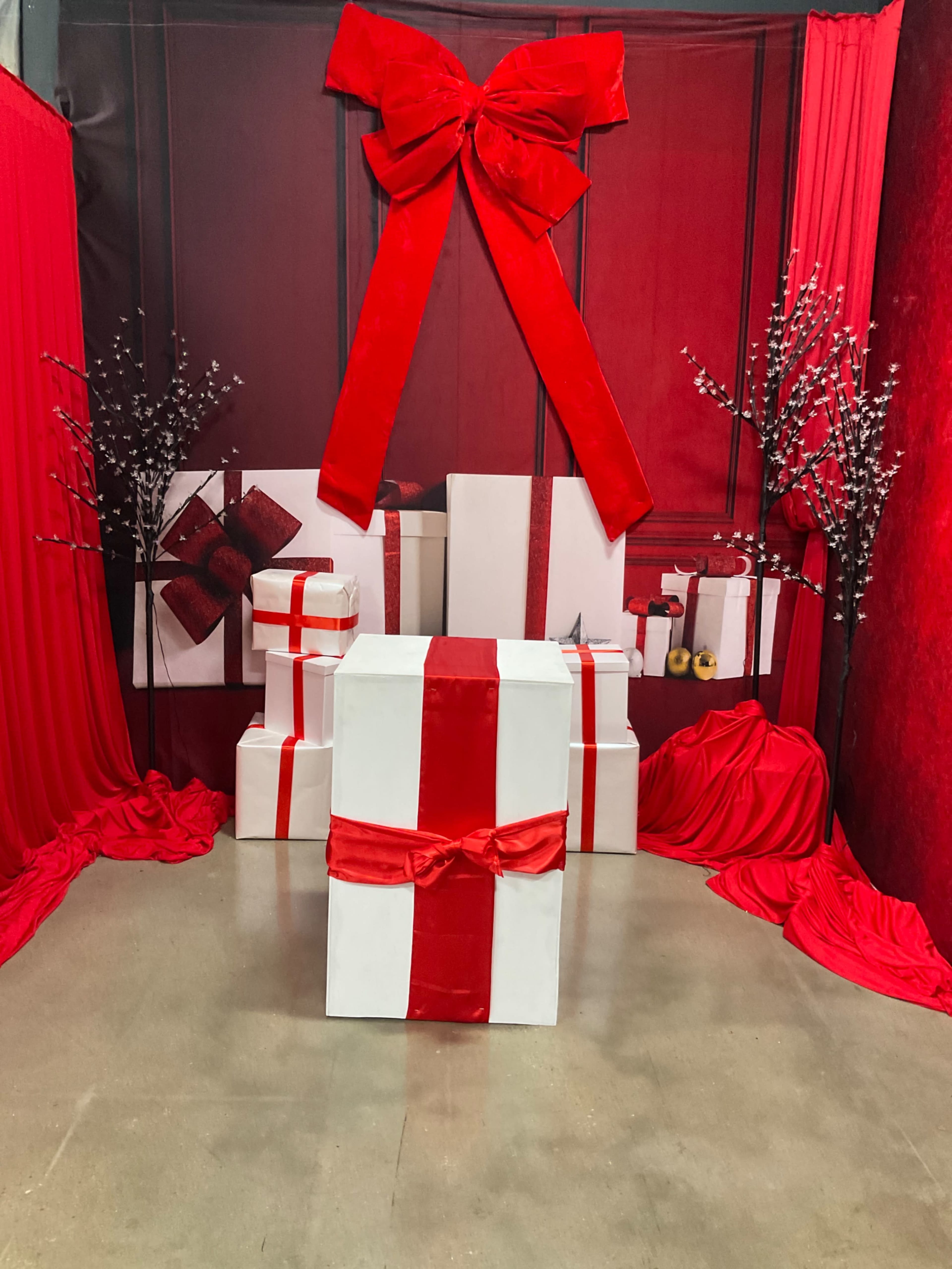 The image features a festive display with large white gift boxes wrapped in red ribbons, set against a backdrop of red drapes and decorative branches.