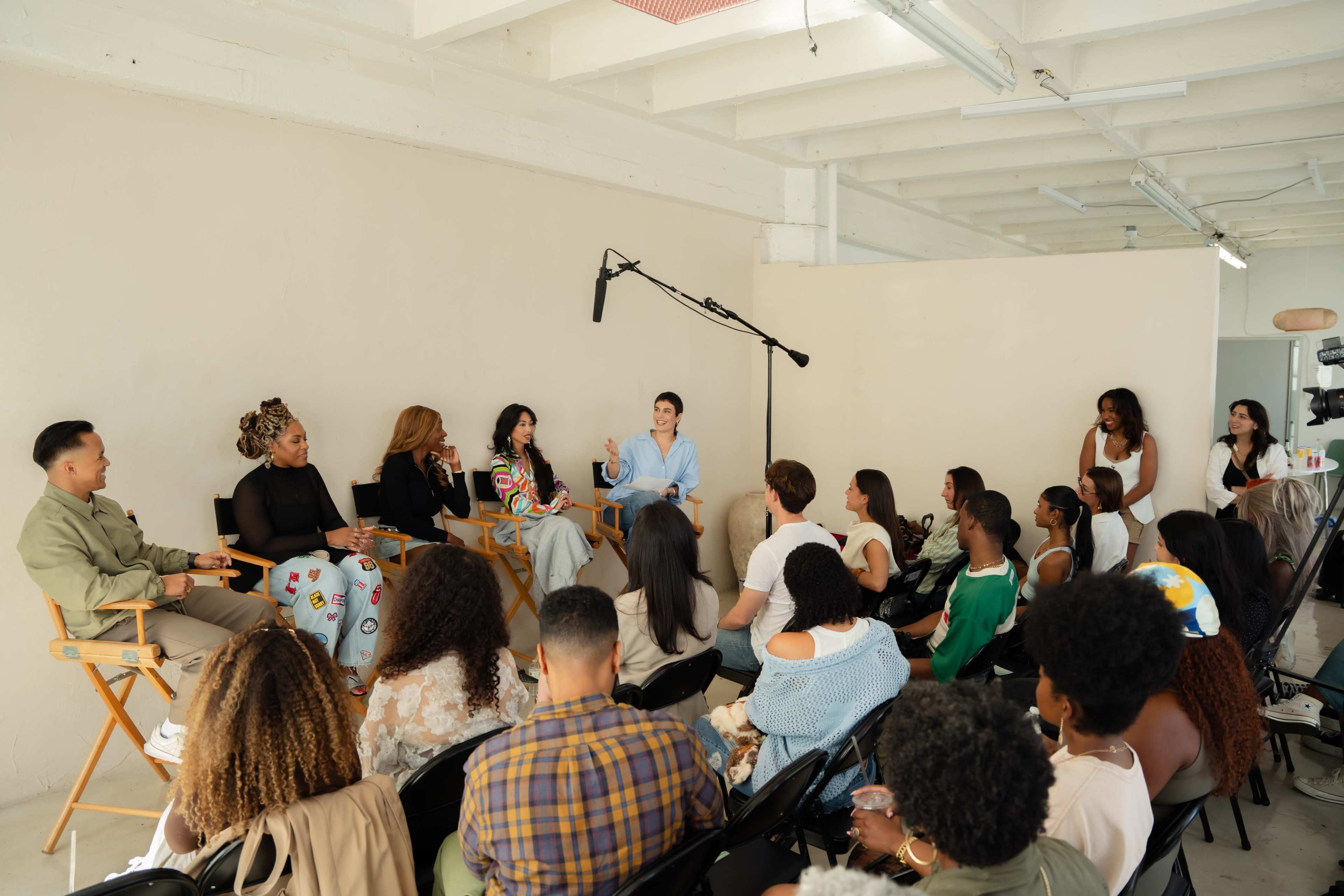 A group of people is sitting in a studio space, attending a panel discussion featuring several speakers seated on directors' chairs.
