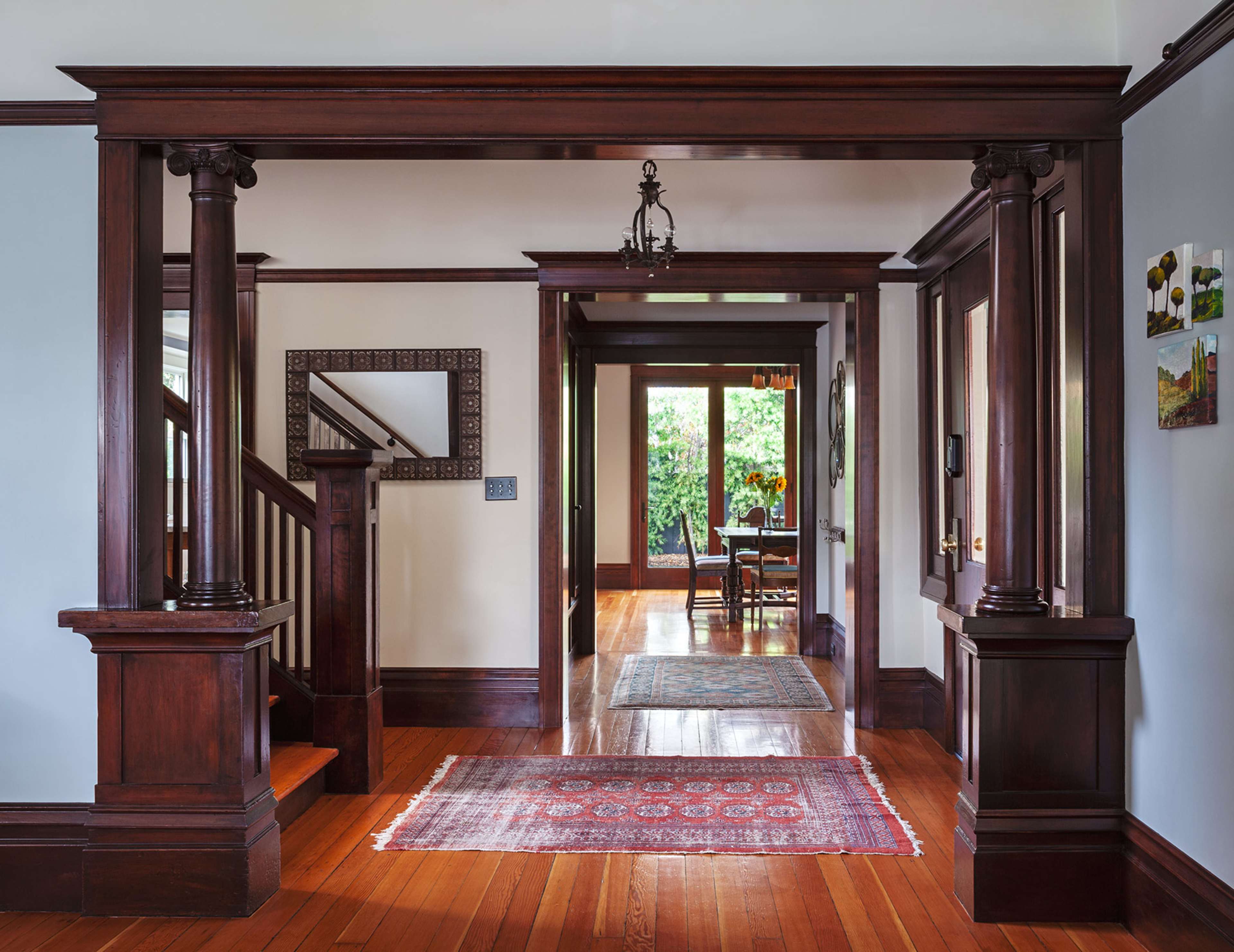 The image shows a spacious entryway with wooden columns, a staircase on the left, and a view into a dining area through an open doorway.