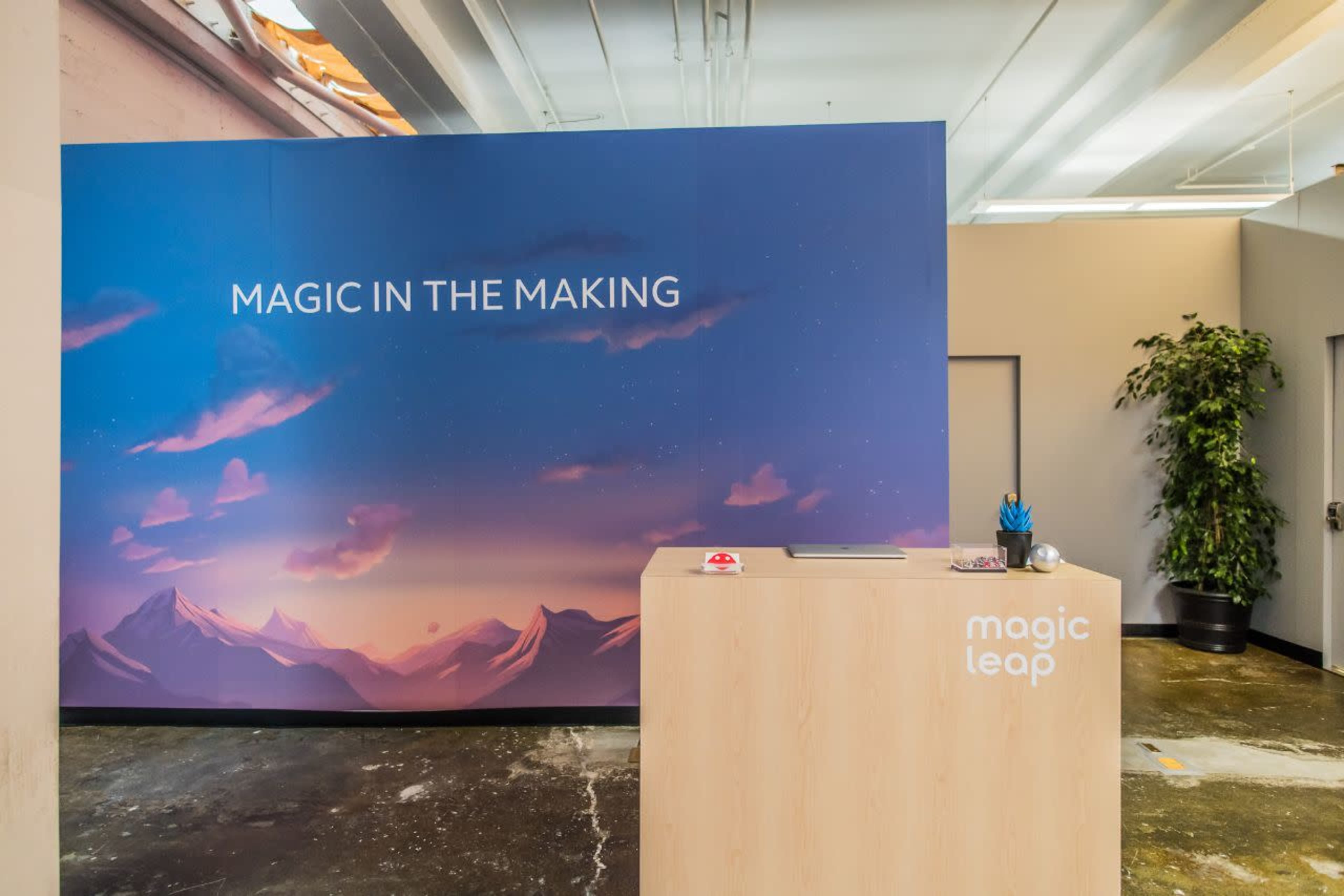 A reception area with a wooden desk and a large backdrop featuring the text "MAGIC IN THE MAKING" against a scenic mountain landscape.