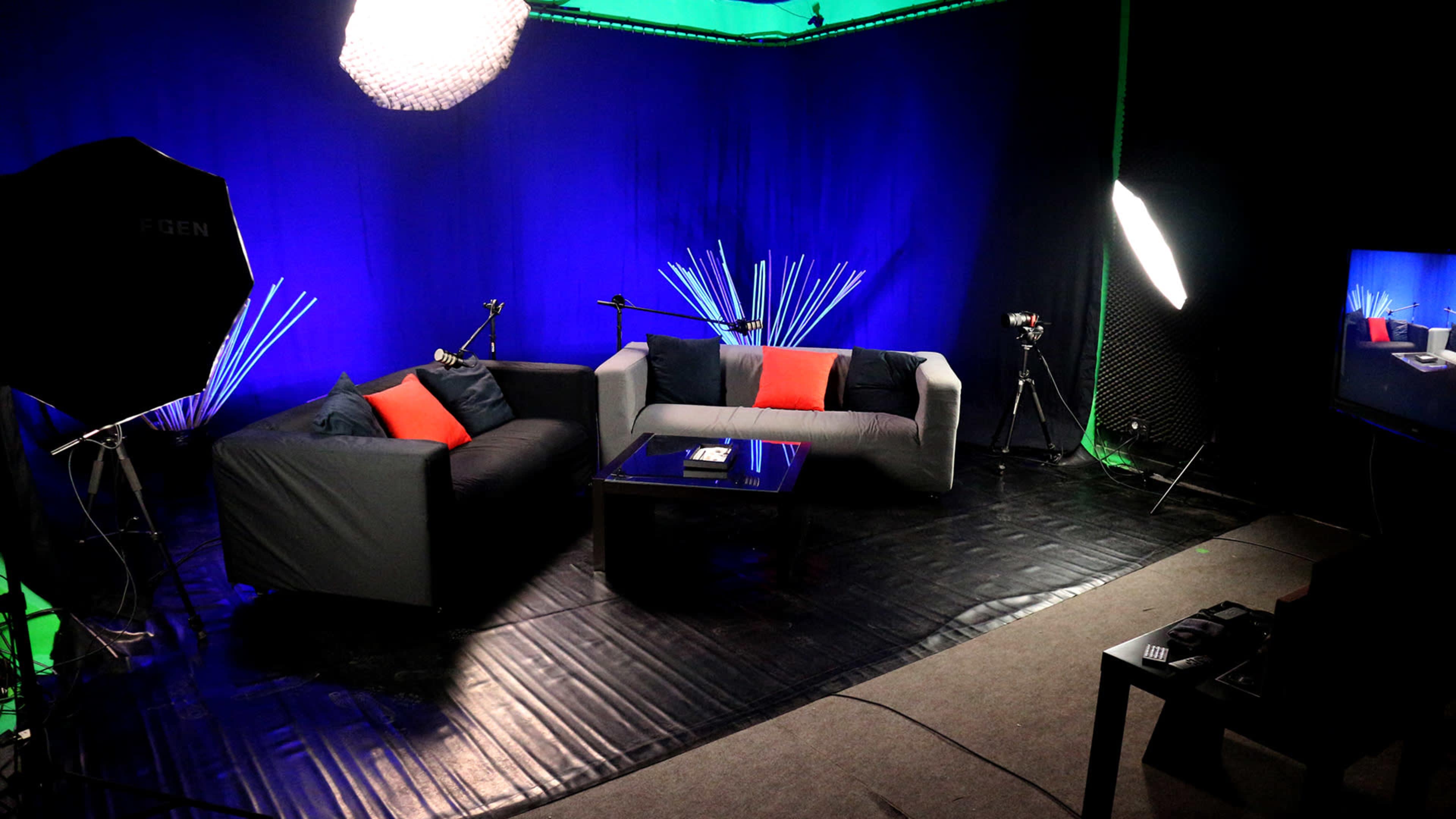 The image depicts a modern studio setting with two couches, a glass coffee table, and various lighting equipment against a backdrop of blue fabric.