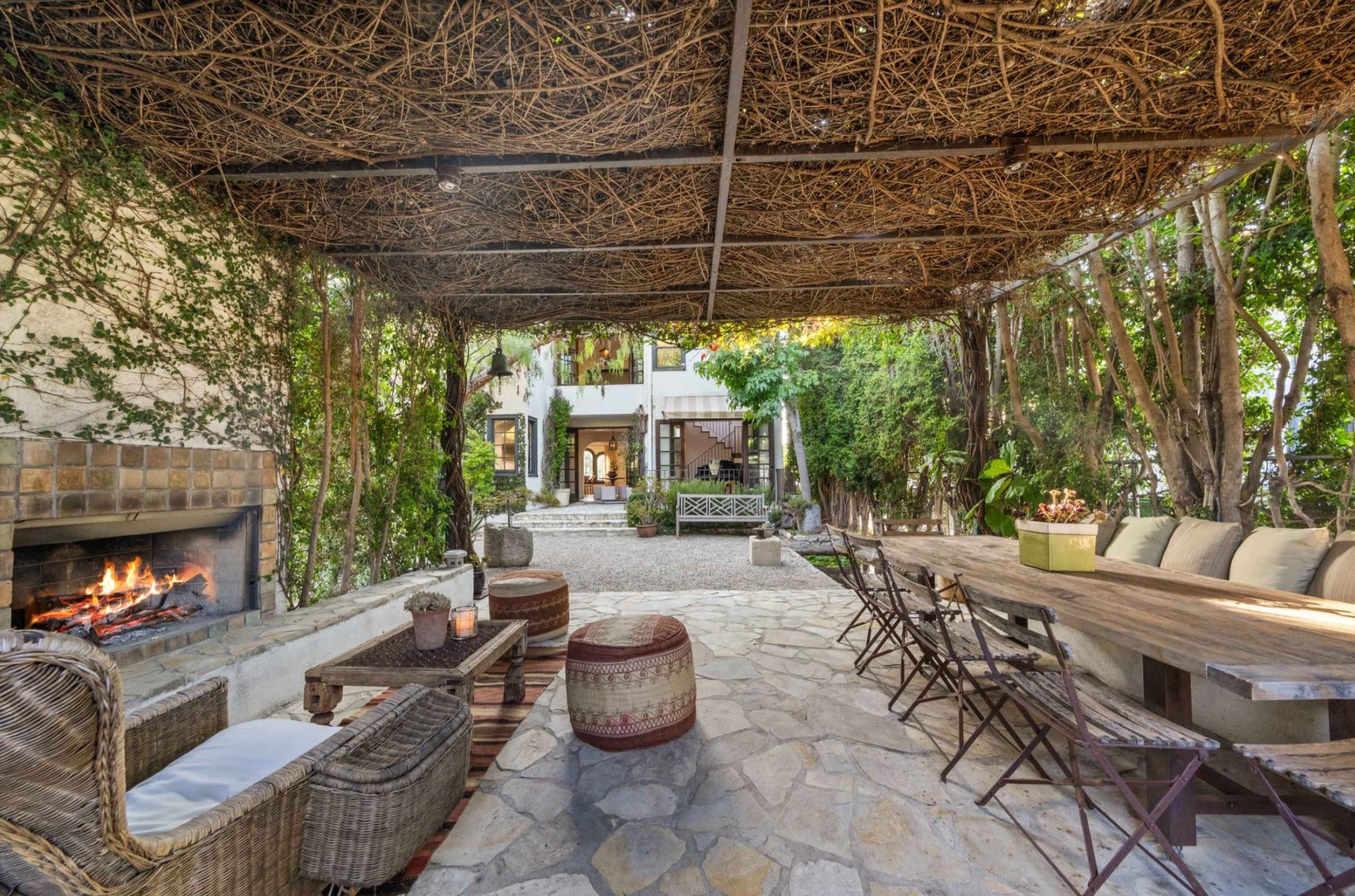 A stone-paved outdoor space features a shaded seating area with a wooden table, surrounded by greenery and a fireplace.