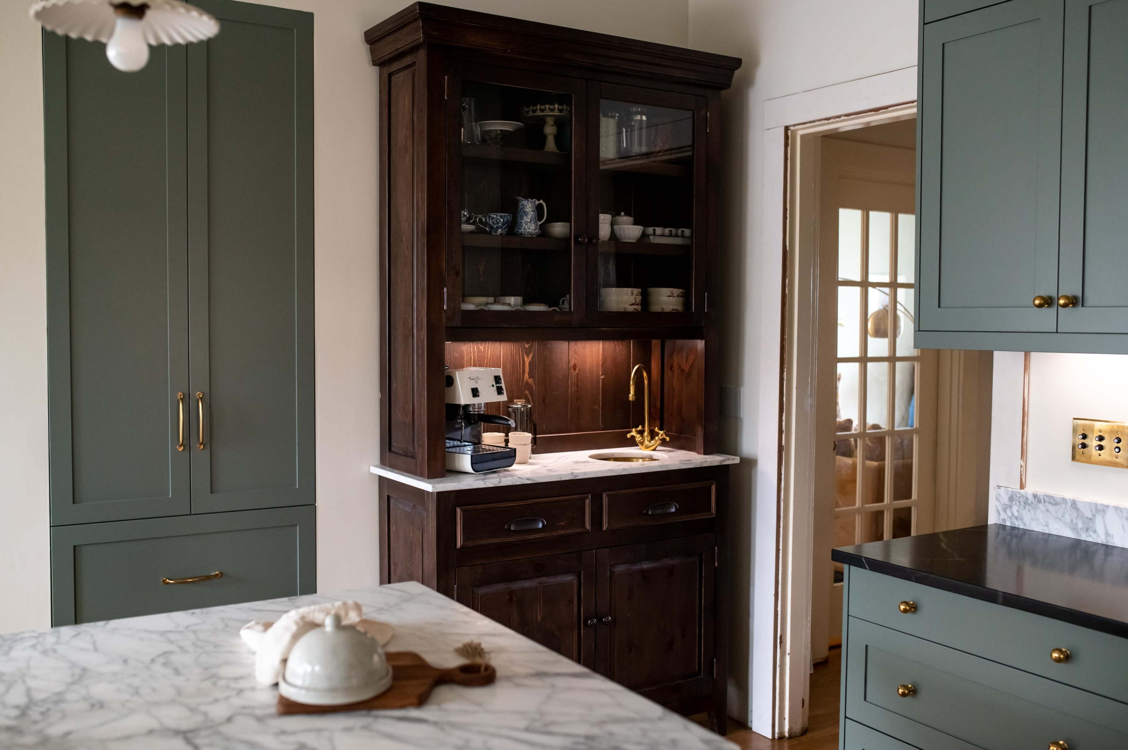 The image features a kitchen area with a dark wooden cabinet displaying dishware, a coffee machine, and a gold faucet, set against green cabinetry and a marble countertop.