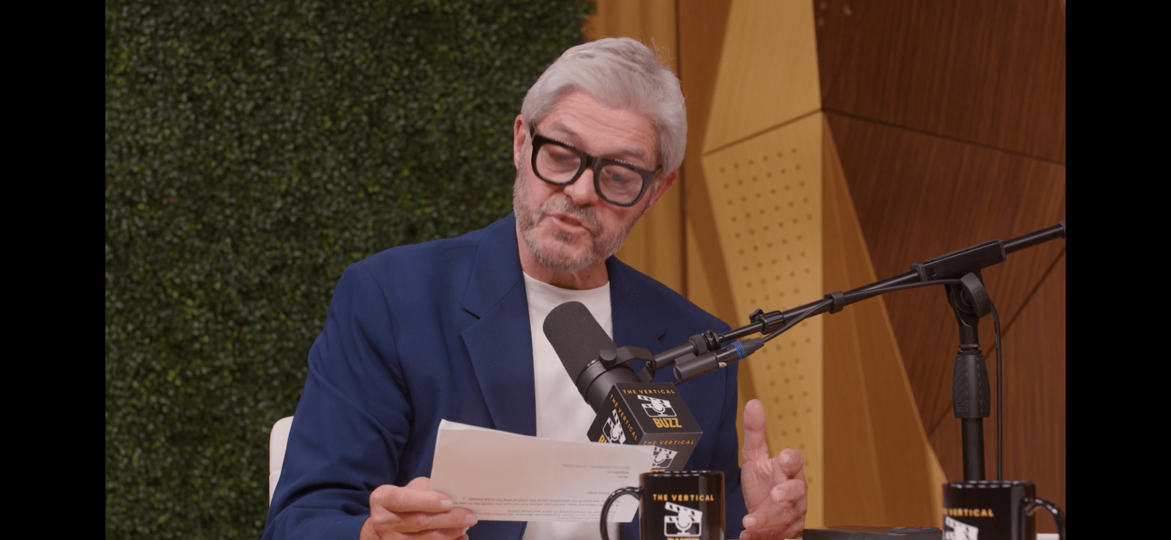 A man with gray hair and glasses reads from a paper while seated at a table with microphones and coffee cups.