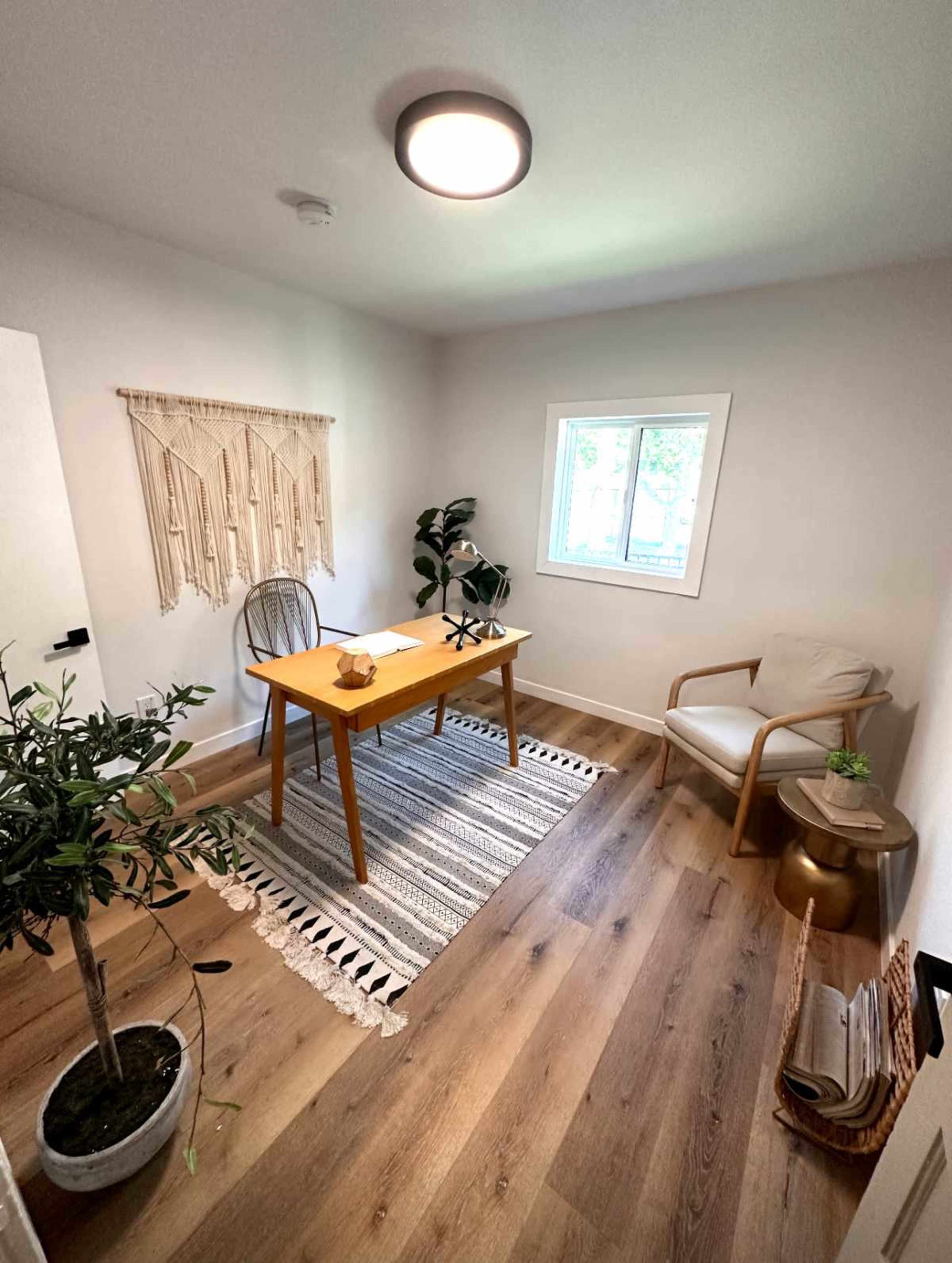 The image shows a small, well-lit workspace featuring a wooden table with a chair, a cozy armchair, a potted plant, and decorative wall hanging.